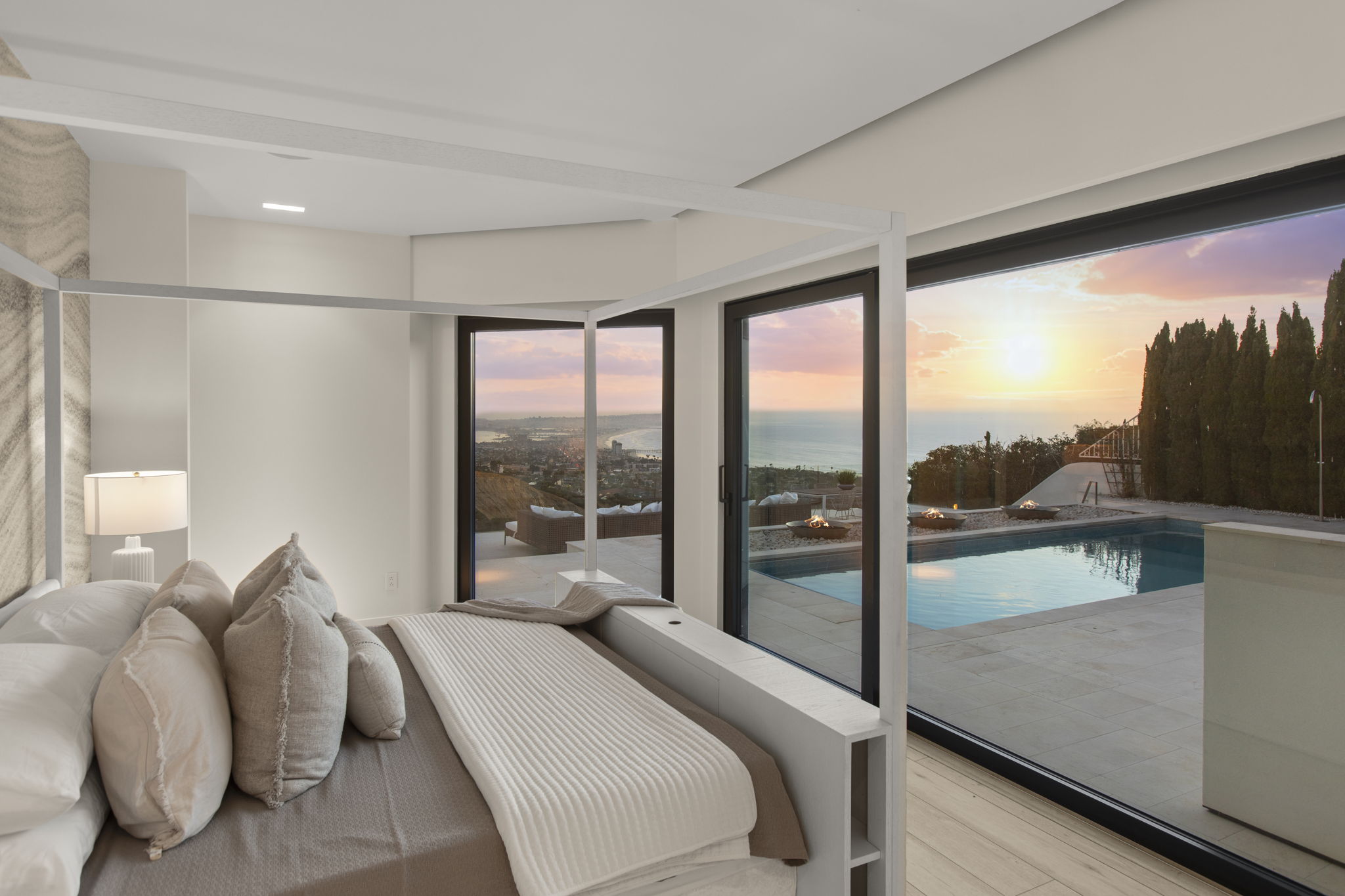 This luxurious primary bedroom features a modern canopy bed frame and a minimalist design aesthetic with neutral tones. A floor-to-ceiling glass wall seamlessly connects the interior space to an outdoor patio, infinity pool, and breathtaking sunset views over the coastal horizon. The sophisticated layout maximizes the connection between indoor comfort and elite outdoor living.