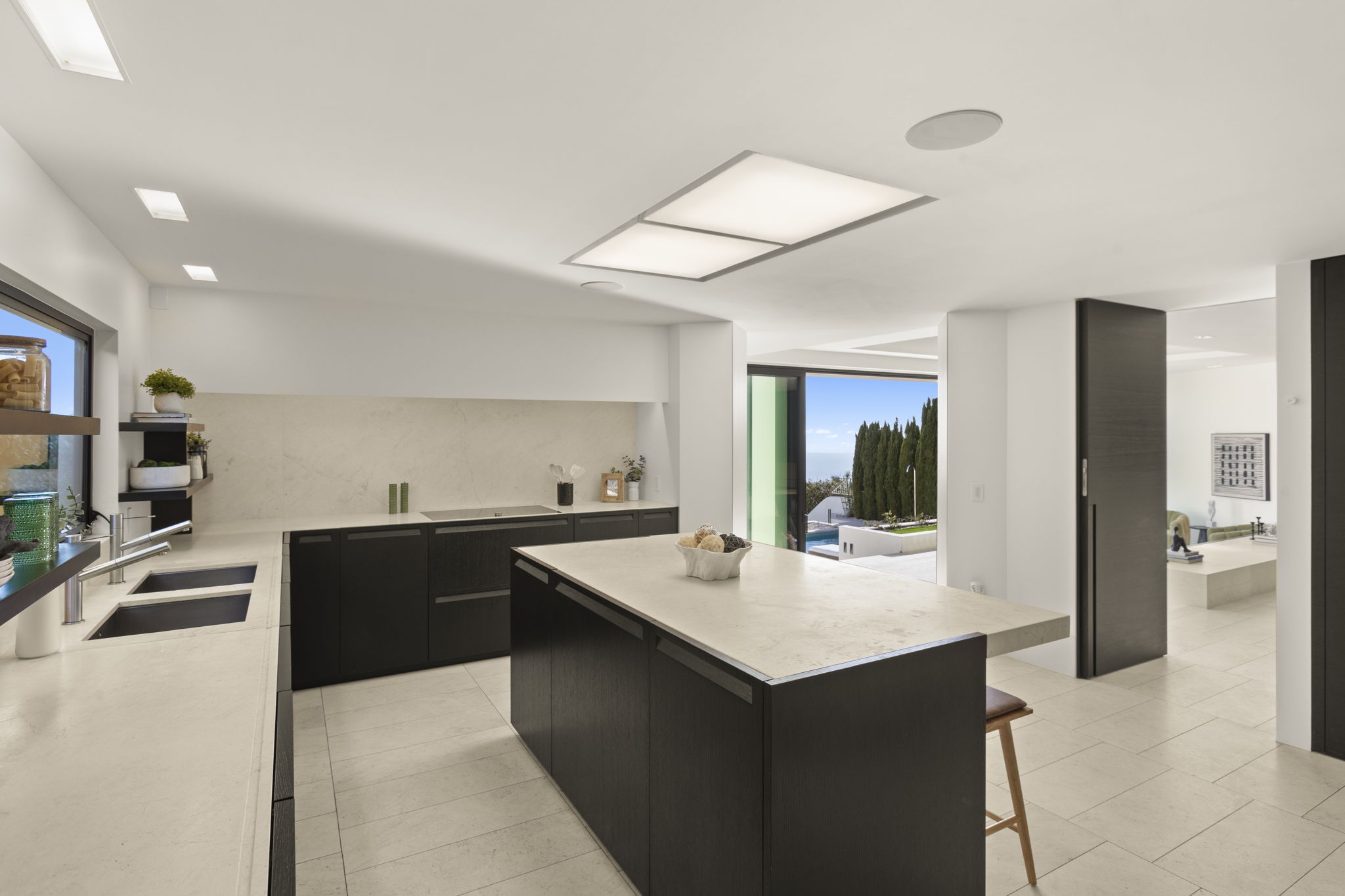 This sleek, contemporary kitchen features dark wood cabinetry, a large central island with stone-look countertops, and integrated high-end appliances. The minimalist aesthetic is complemented by clean lines, a neutral color palette, and an open layout that seamlessly connects to an outdoor view of a pool area. The space feels both sophisticated and functional, highlighting premium finishes and modern architectural lighting.