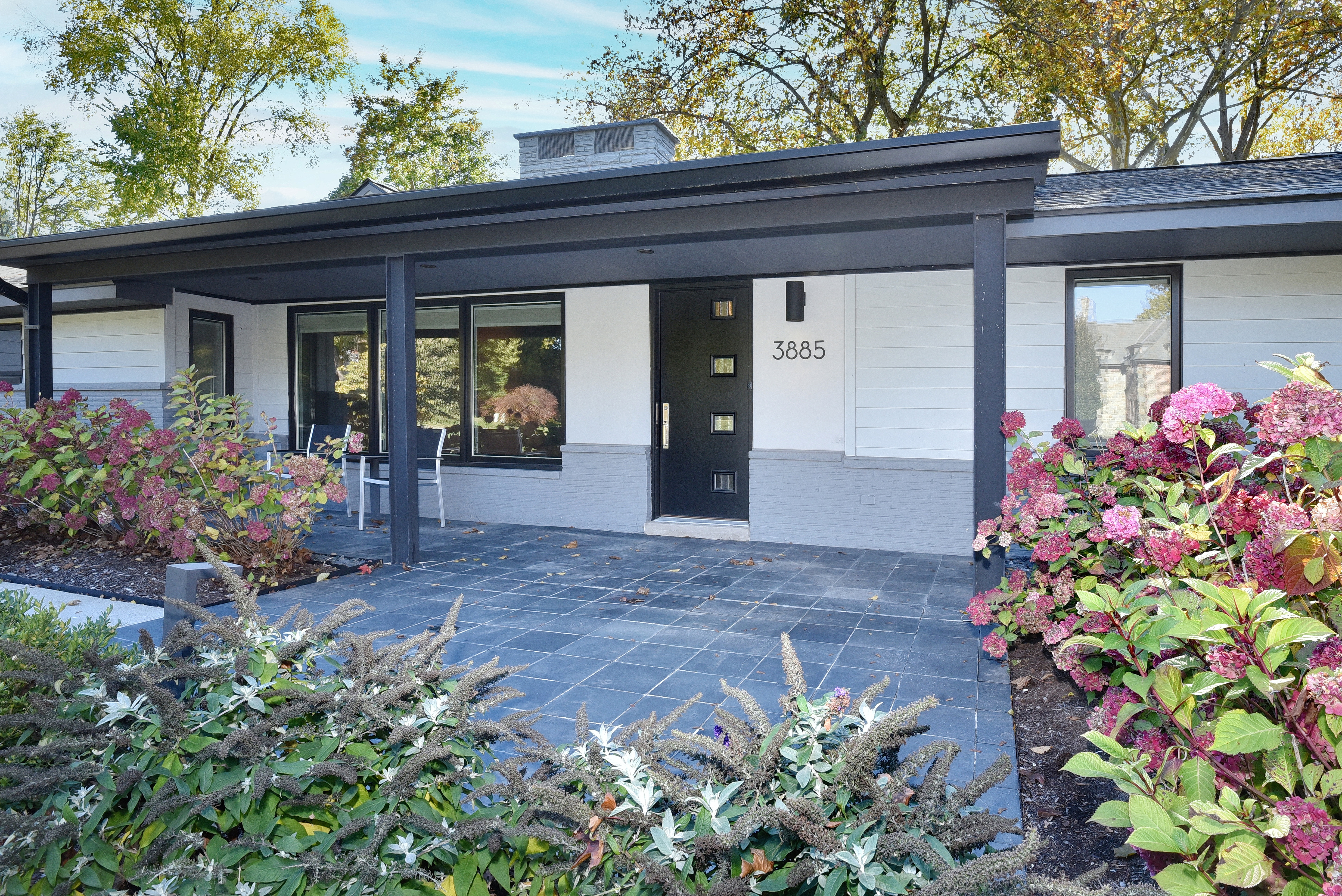 The image showcases the entryway of a modern home, featuring a dark-colored front door with unique square windows and the house number "3885" displayed above. The facade combines white siding with a gray brick base, complemented by dark trim and a flat roof overhang. The entryway is adorned with lush landscaping, including hydrangeas and other greenery, adding to the home's curb appeal.