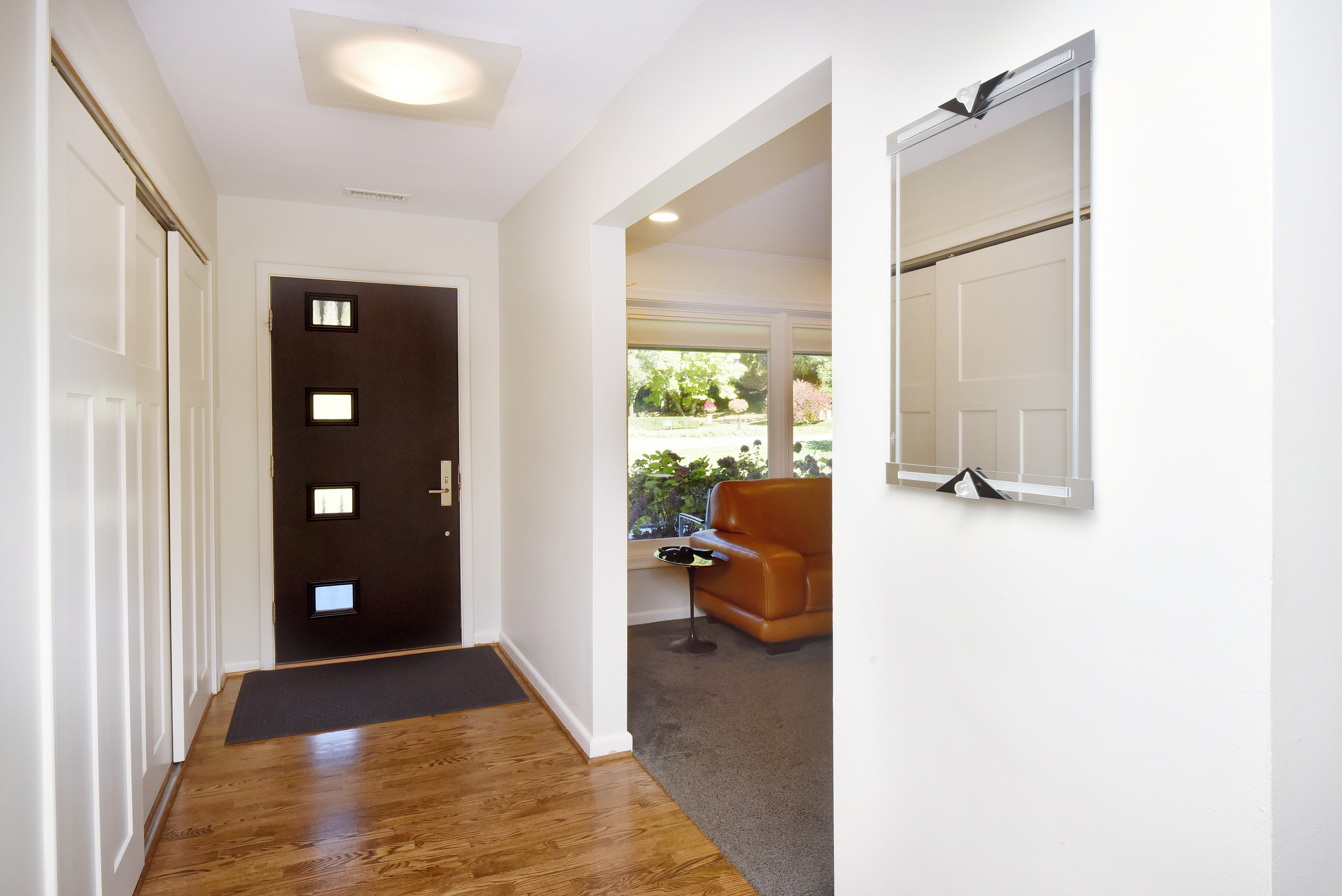 This interior shot showcases a well-lit hallway with hardwood flooring and white walls, leading to a living area. A modern mirror hangs on the wall, reflecting the hallway's features, while a dark-colored front door with square windows adds a contemporary touch. The space feels clean and inviting, highlighting the home's modern aesthetic.
