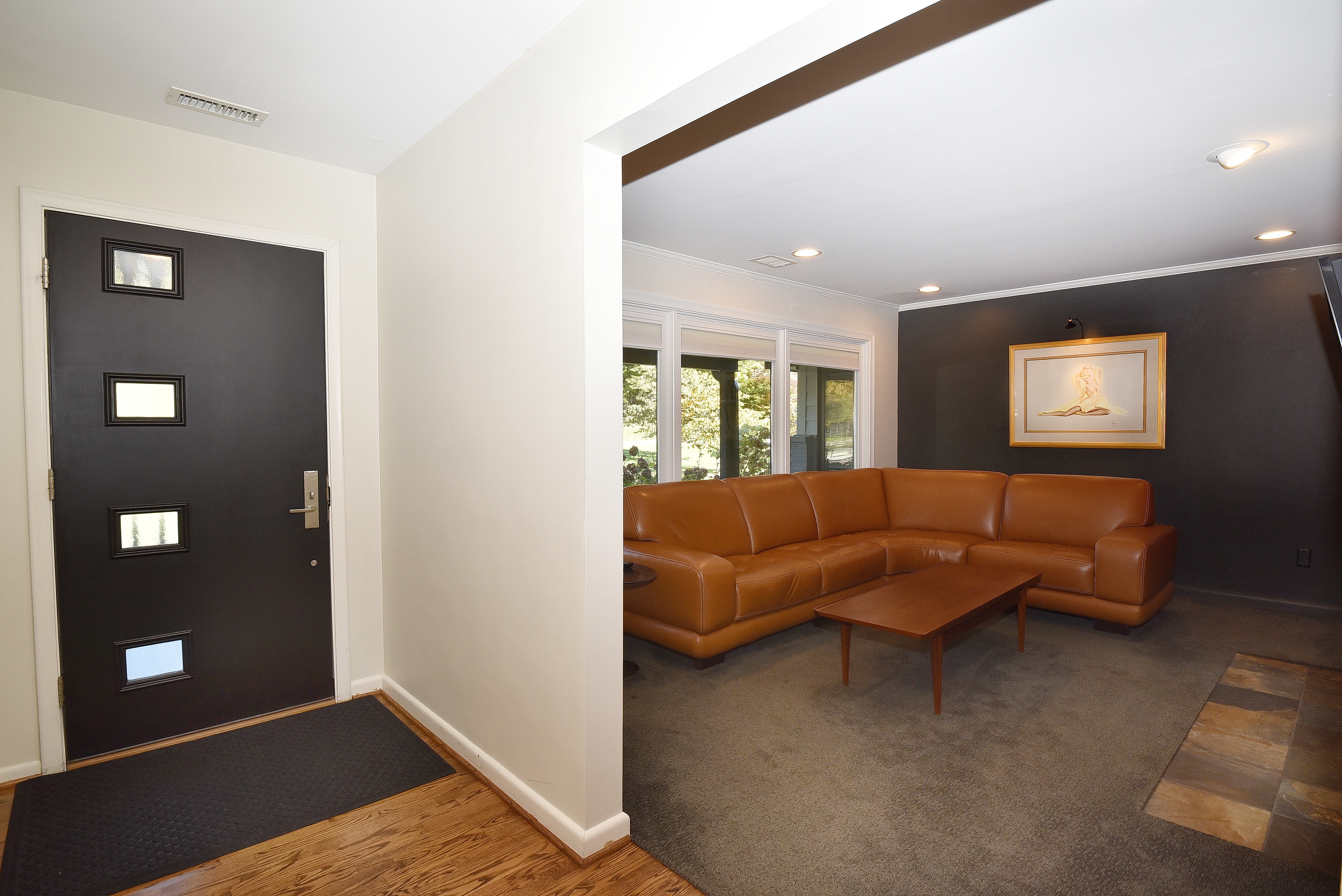 This interior shot showcases a living room with a modern aesthetic. A large, brown leather sectional sofa and a wooden coffee table anchor the space, set upon a neutral-toned carpet. The room features a dark accent wall with a framed artwork, complemented by recessed lighting and a view through a window, creating a warm and inviting atmosphere.