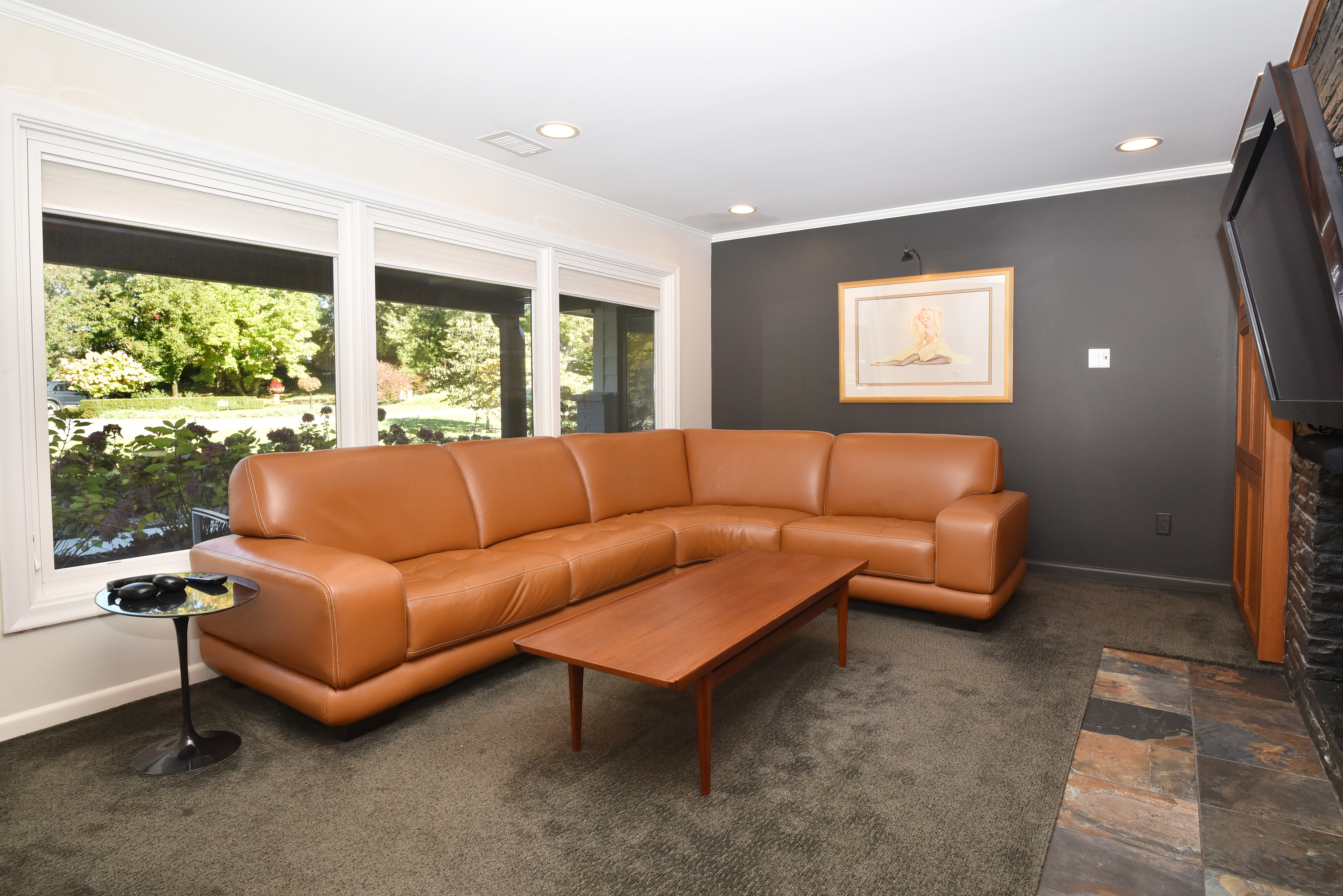 This living room features a large, caramel-colored leather sectional sofa, a wooden coffee table, and a black side table with a glass top. The room is well-lit by natural light streaming through a large window, and the walls are painted in a combination of white and dark gray, with an artwork hanging on the dark gray wall. The flooring is carpeted, and there is a fireplace with a stone surround on the right side of the image.
