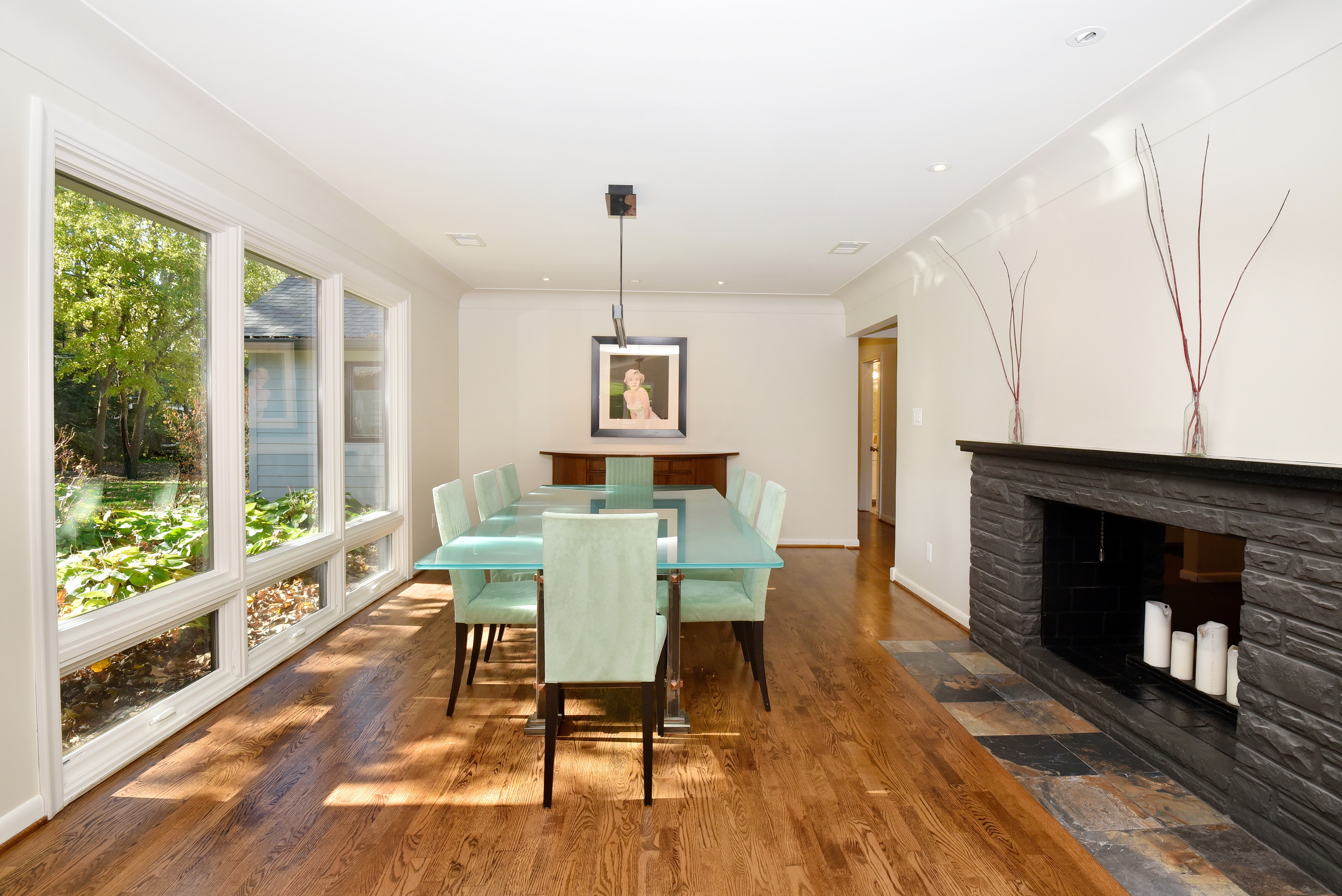 This dining room features a modern aesthetic with a glass-top dining table surrounded by light green chairs. A large window provides ample natural light, highlighting the hardwood floors. A fireplace with a stone surround adds a touch of warmth and character to the space.