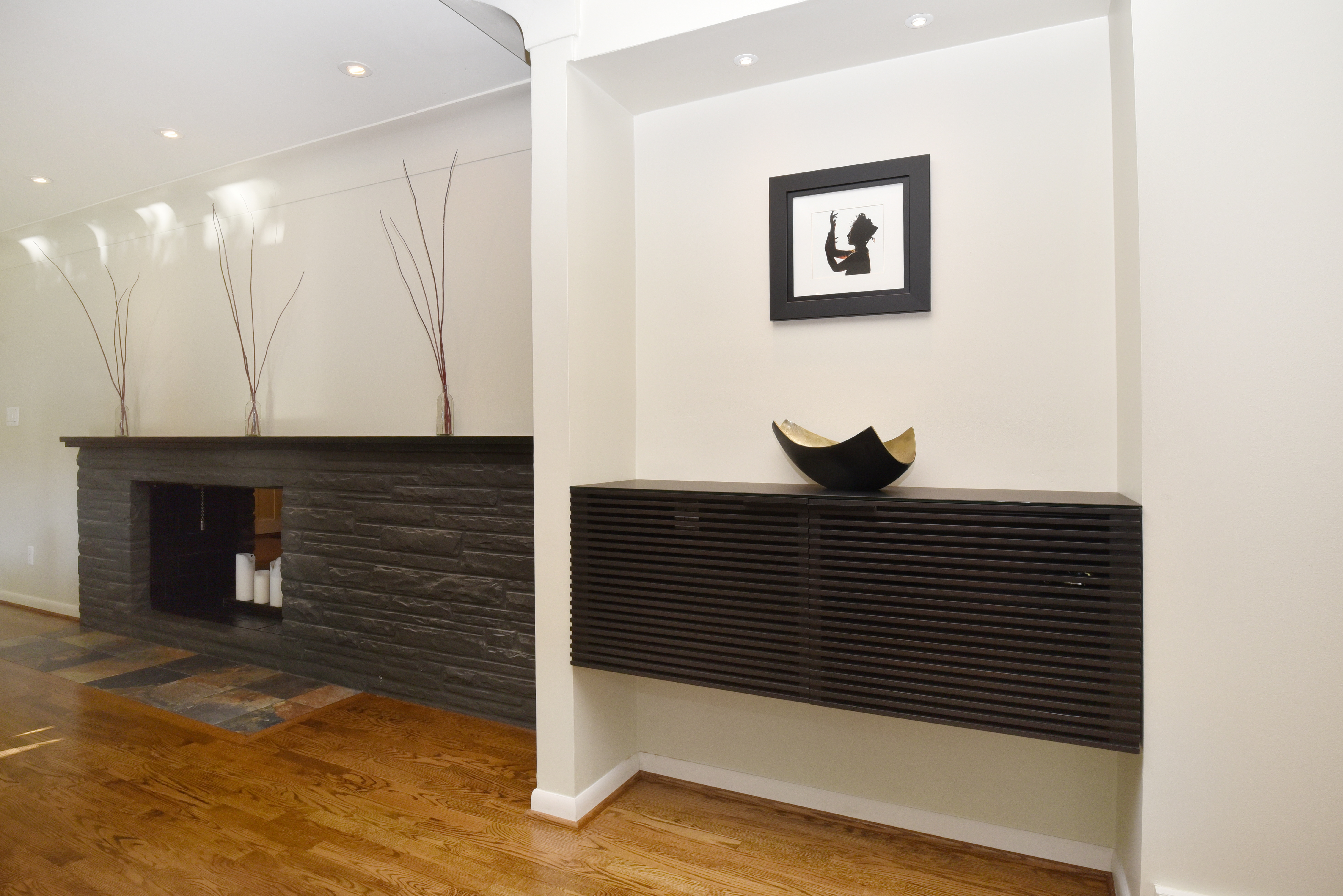The image showcases a modern living room with a sleek fireplace featuring a dark stone facade and a floating dark wood cabinet with a black and gold decorative bowl. A framed artwork hangs above the cabinet, and the hardwood floors add warmth to the space. The room's design is minimalist and sophisticated.
