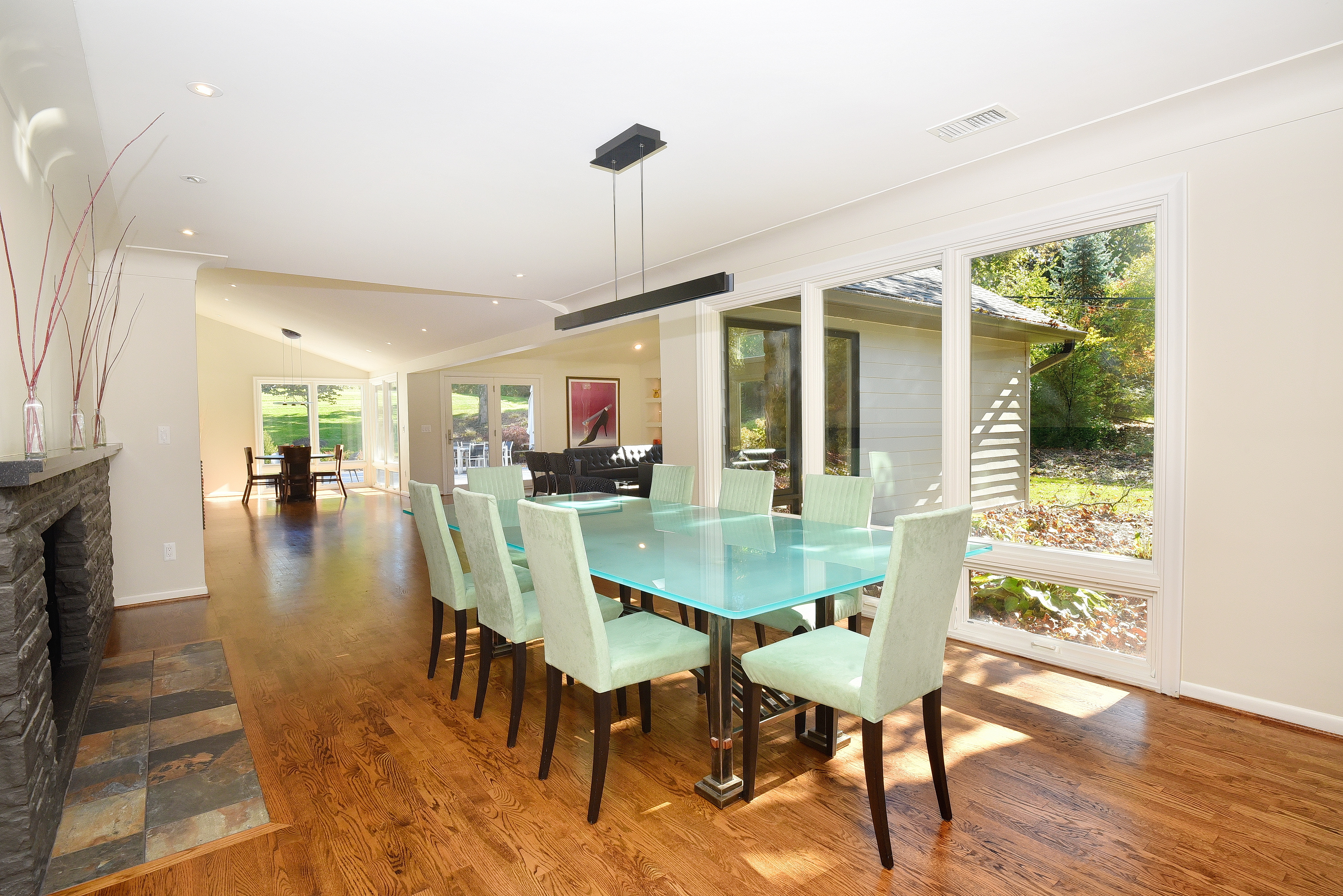 This is an interior shot of a dining room featuring a glass-top dining table with seating for eight. The room has hardwood floors and a large window offering natural light and a view of the outdoors. A modern linear chandelier hangs above the table, and a fireplace is visible on the left, adding a touch of warmth to the space.
