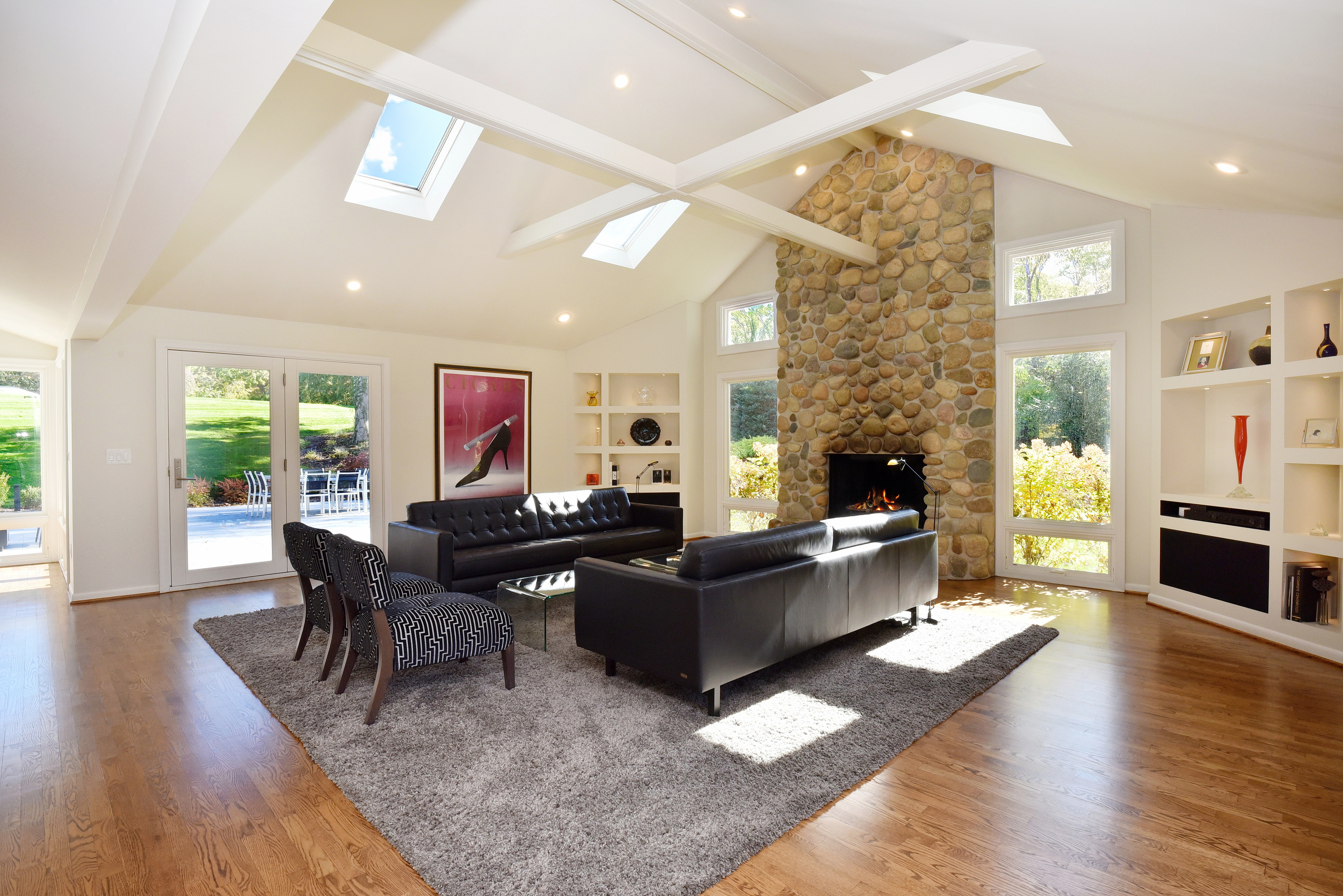 This is a spacious living room featuring a high vaulted ceiling with skylights and exposed beams. A stone fireplace serves as a focal point, complemented by two black leather sofas and patterned armchairs arranged on a large gray rug. The room is bright and airy, with natural light streaming in through large windows and glass doors leading to the outside.