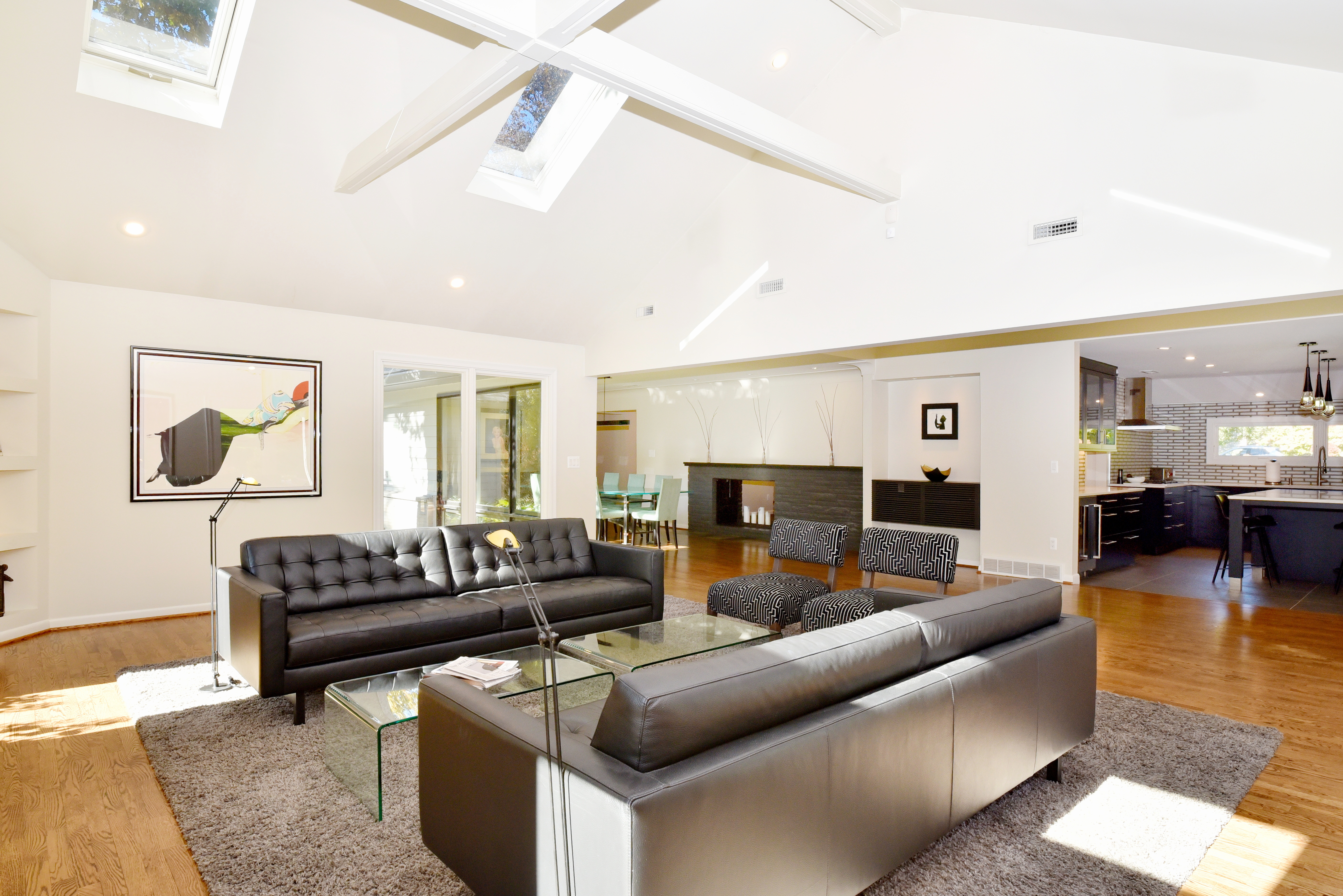This is a spacious living room featuring two black leather sofas, a glass coffee table, and a gray rug. The room has a high, vaulted ceiling with skylights, and a modern fireplace is visible in the background. The open floor plan connects to the dining area and kitchen, creating a bright and inviting space.
