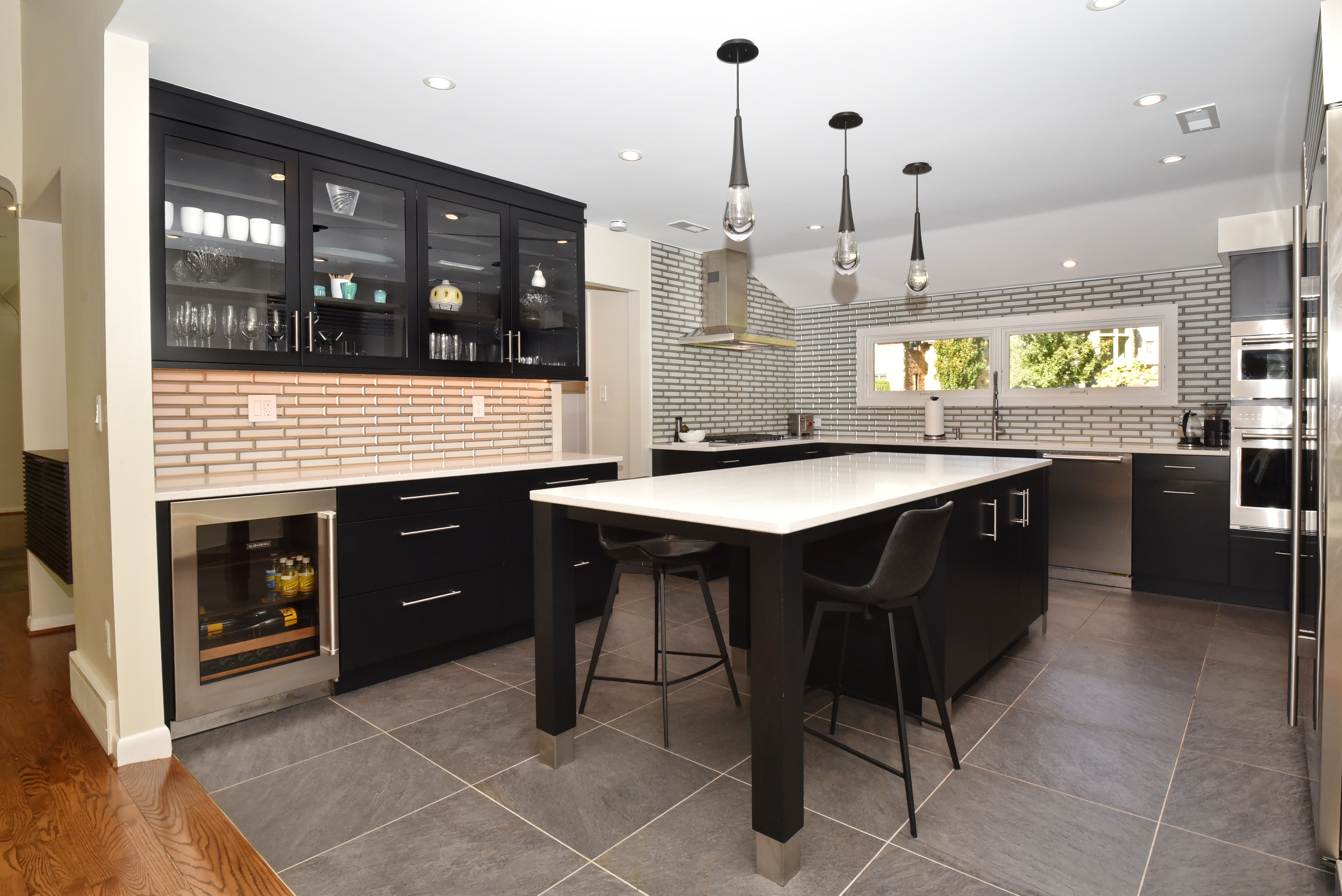 This is a modern kitchen featuring black cabinetry, stainless steel appliances, and a large island with a white countertop. The kitchen has a sleek design with pendant lighting above the island and a wine cooler built into the cabinetry. The backsplash is a light-colored brick pattern, and the flooring is a large gray tile.