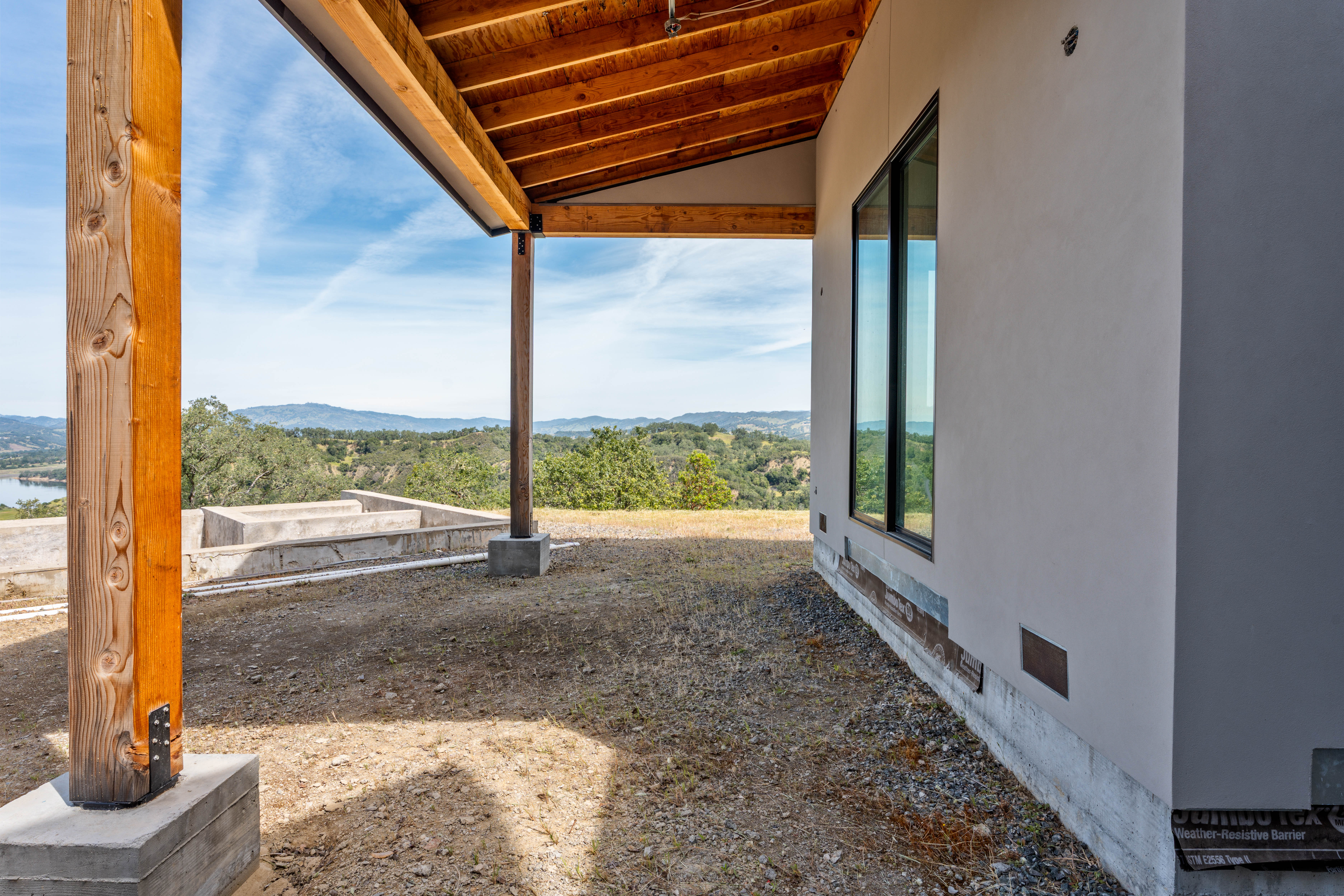 This image captures a covered outdoor patio area under construction, featuring exposed wooden beams and a sturdy timber support post. The space offers an expansive, scenic view of rolling hills and a distant lake, highlighting the property's prime location. The unfinished ground and exposed foundation elements suggest a modern home in the final stages of completion, emphasizing potential for outdoor living.