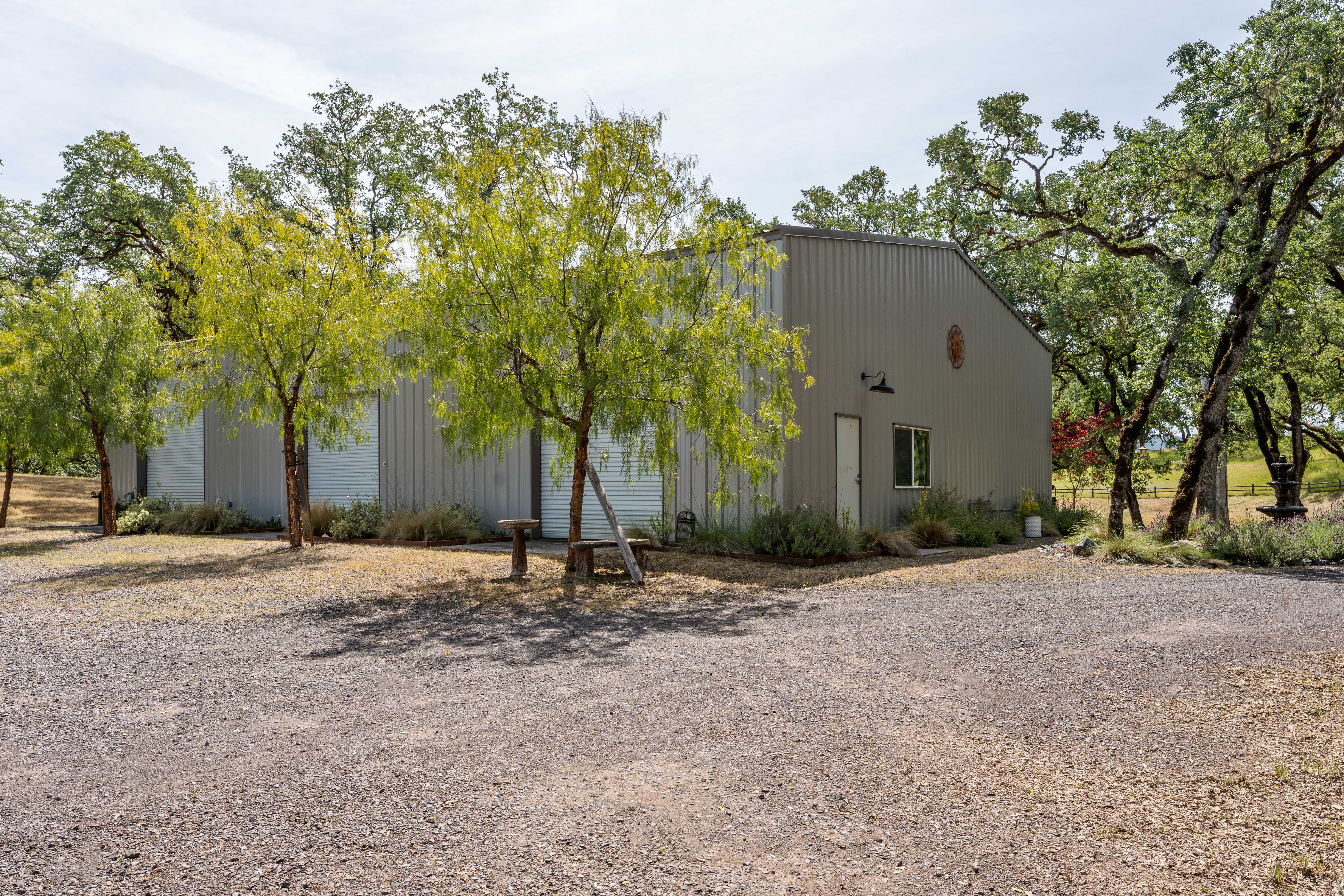 This image showcases a large, metal-sided workshop or garage building situated on a gravel lot, surrounded by mature oak trees. The structure features multiple roll-up doors, a side entry door, and a window, presenting a functional and utilitarian aesthetic. The scene is bathed in bright, natural daylight, emphasizing the spacious outdoor area and the rural, peaceful setting.