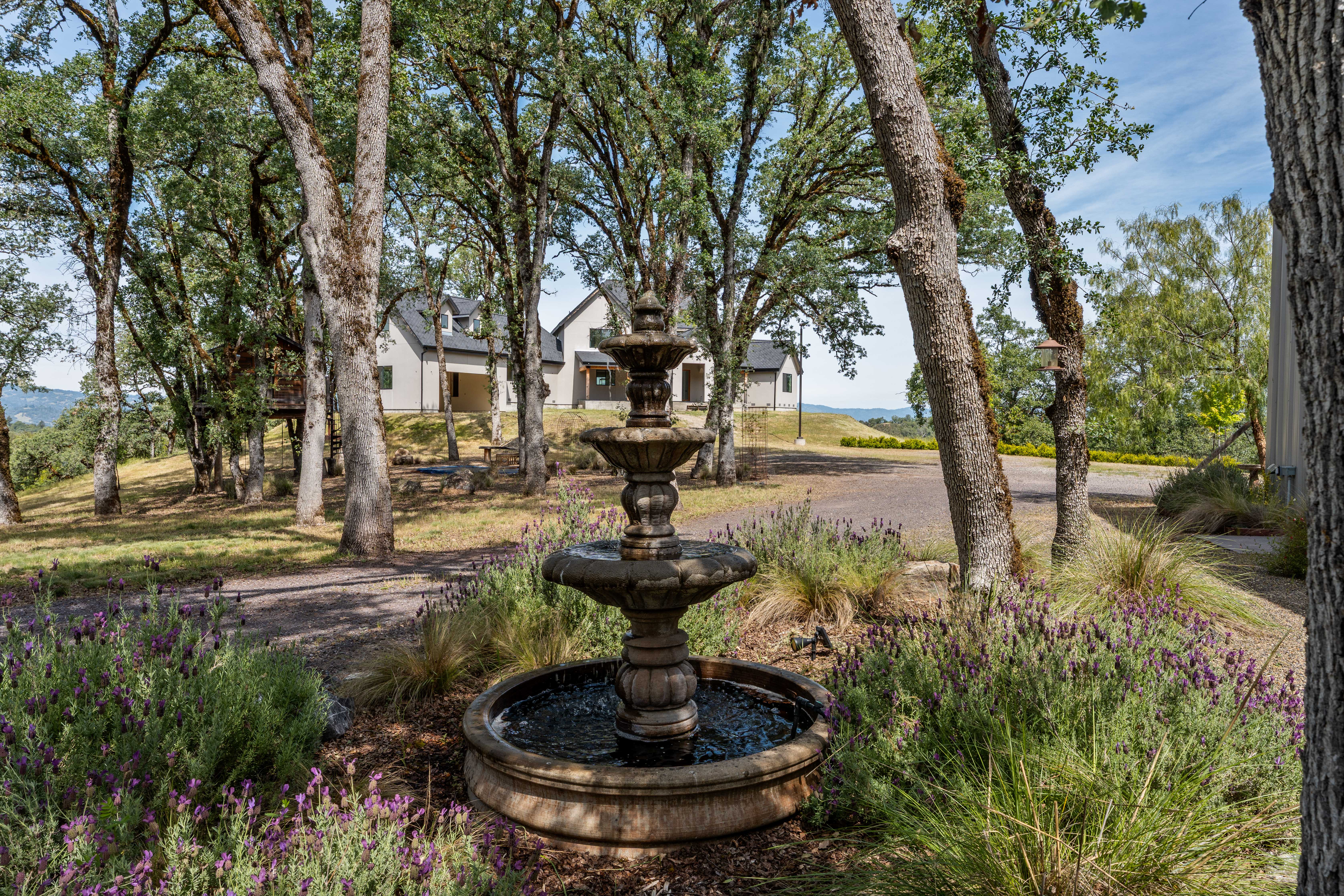 This serene outdoor scene features a classic tiered stone fountain as the focal point, surrounded by lush lavender bushes and mature oak trees. In the background, a modern farmhouse-style residence sits atop a gentle slope, offering a picturesque view of the property's expansive, wooded landscape. The composition uses the fountain to draw the eye toward the home, creating a peaceful and inviting atmosphere.