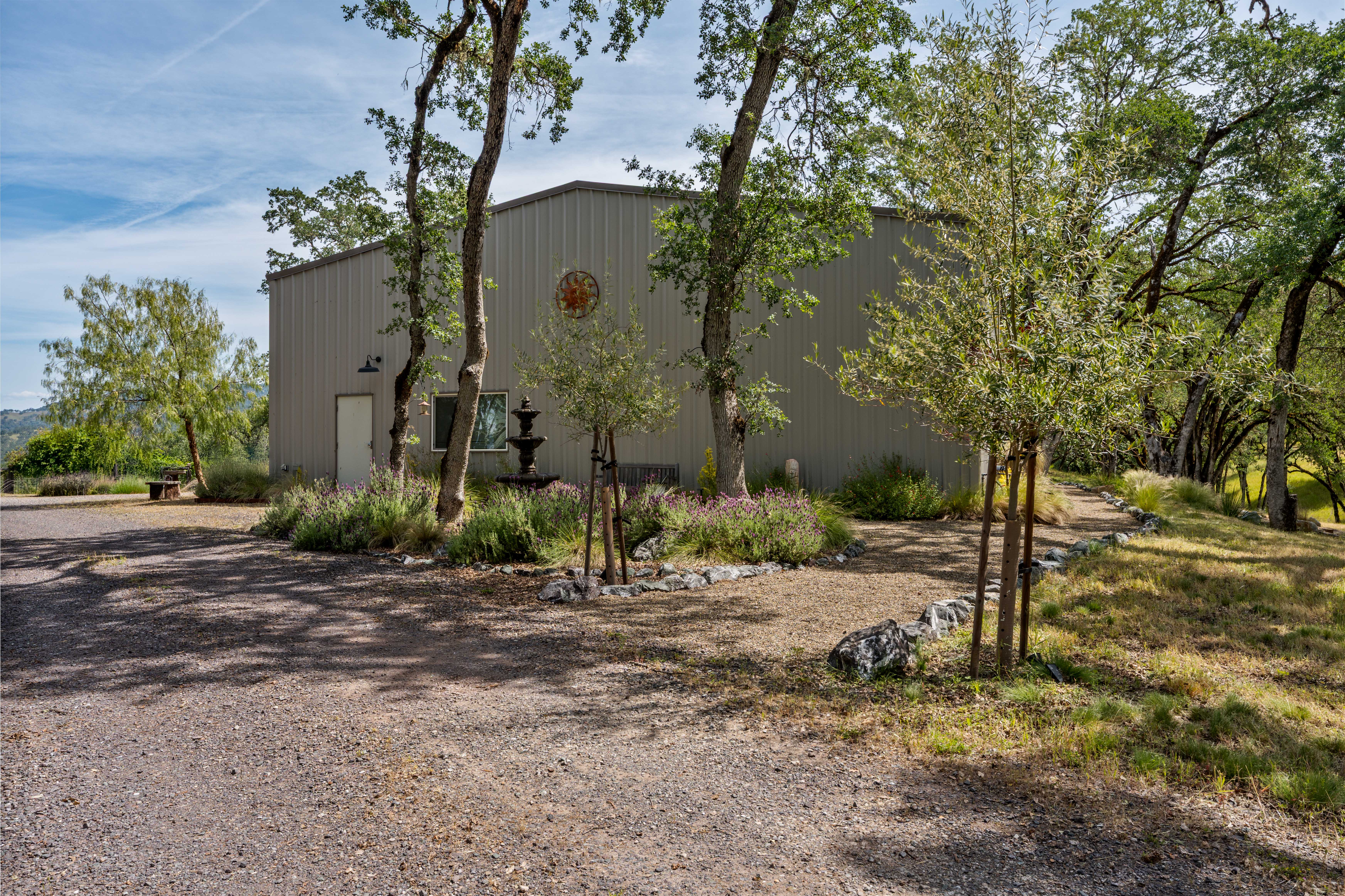 This side view of a metal-sided structure showcases a rustic, rural aesthetic set against a backdrop of mature oak trees and a gravel driveway. A decorative fountain and a small garden bed with lavender add a touch of charm to the building's exterior, while the surrounding landscape emphasizes a peaceful, countryside setting. The perspective captures the building's scale and its integration into the natural environment.