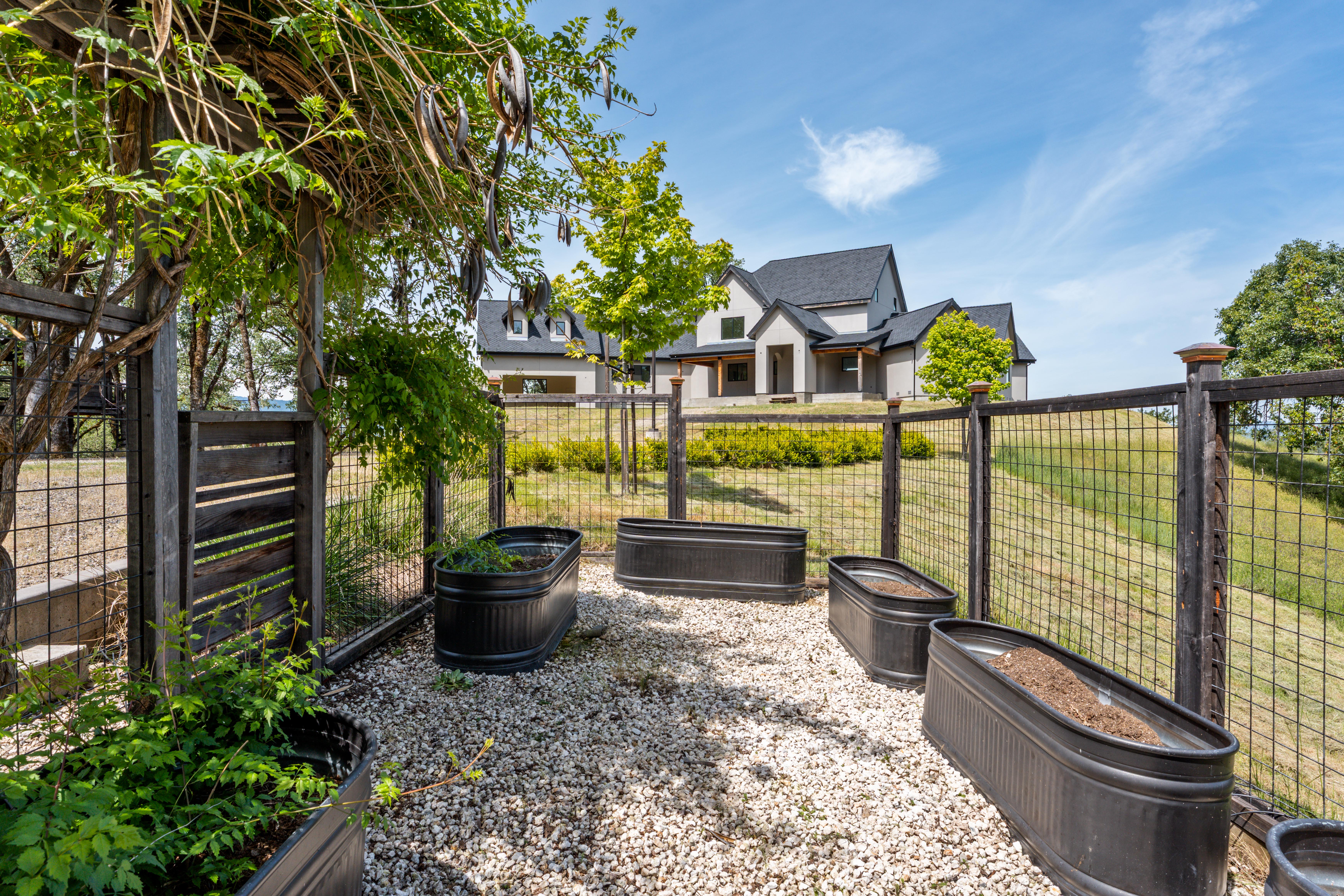This image captures a well-organized garden area featuring several black metal raised planter beds set upon a gravel base. The space is enclosed by a rustic wooden and wire mesh fence, with a lush vine-covered trellis providing shade in the foreground. In the background, a modern, multi-gabled house sits prominently on a gentle hill, creating a serene and picturesque residential setting.