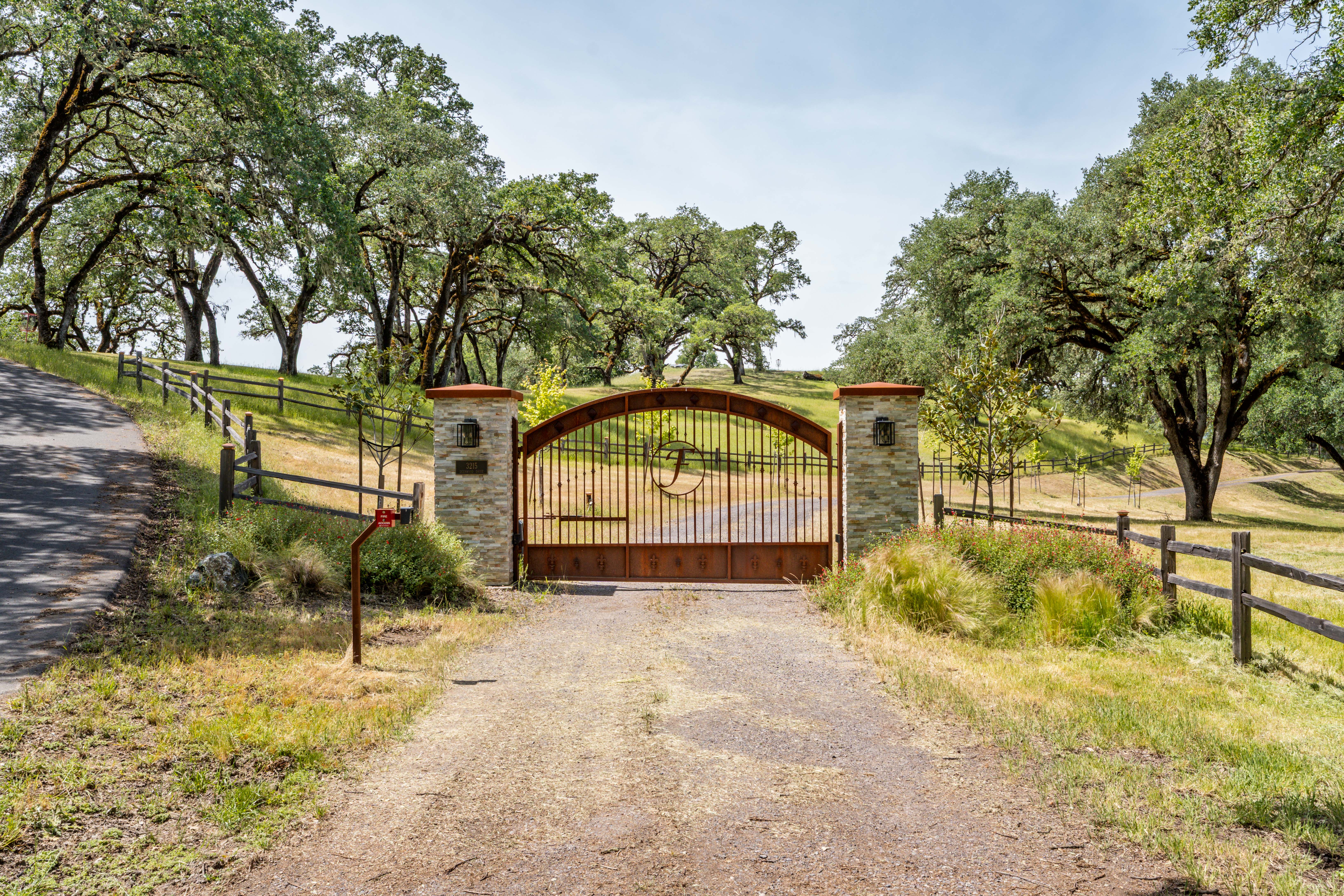 This image captures the grand gated entrance to a private estate, featuring a rustic, arched metal gate flanked by stone pillars. The driveway leads through a picturesque landscape of rolling hills and mature oak trees, creating a sense of seclusion and prestige. The scene is framed by wooden fencing and natural, sun-drenched vegetation, offering a welcoming and secure first impression of the property.