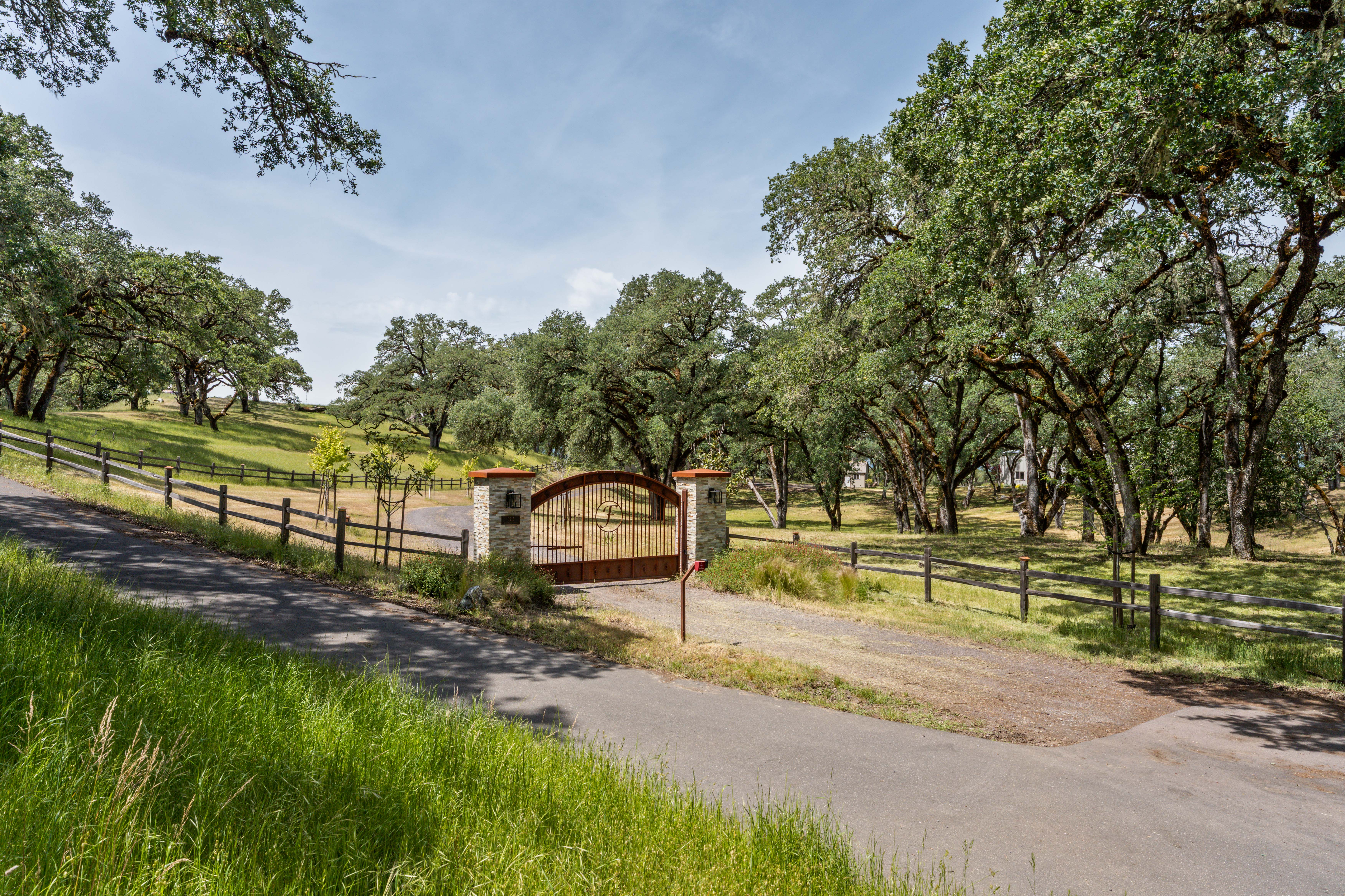 This image captures the grand, gated entrance to a sprawling rural estate, featuring a rustic metal gate flanked by stone pillars. A paved driveway leads through the gate, framed by expansive grassy hills and mature oak trees that create a serene, private atmosphere. The scene is shot from a low-angle perspective, emphasizing the welcoming and secure nature of the property's approach.