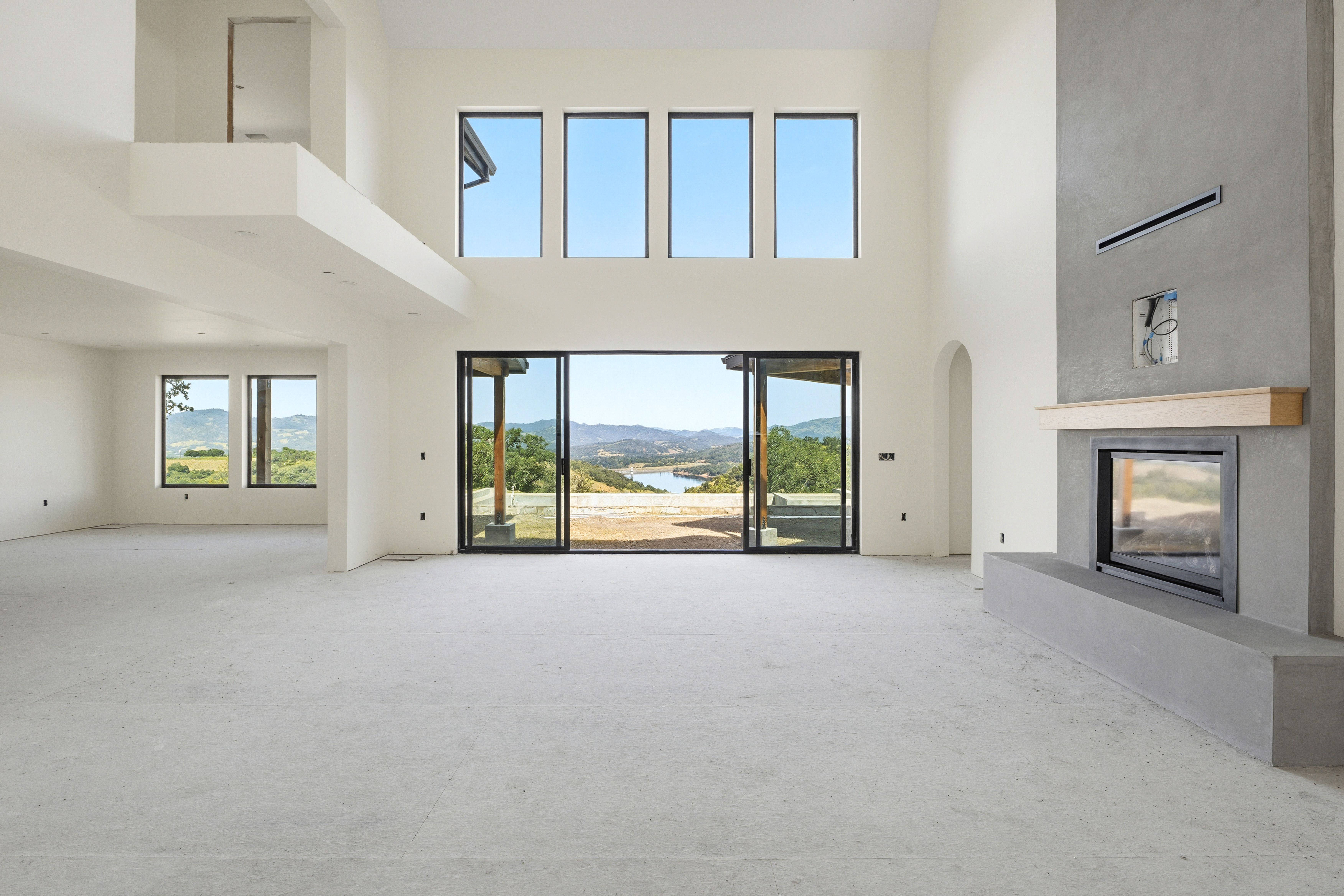 This expansive, double-height living room features a minimalist aesthetic with unfinished concrete flooring and crisp white walls. A striking floor-to-ceiling gray concrete fireplace serves as the focal point, while massive windows and sliding glass doors frame a breathtaking panoramic view of the surrounding hills and lake. The open-concept design creates a bright, airy atmosphere that emphasizes the home's connection to the natural landscape.