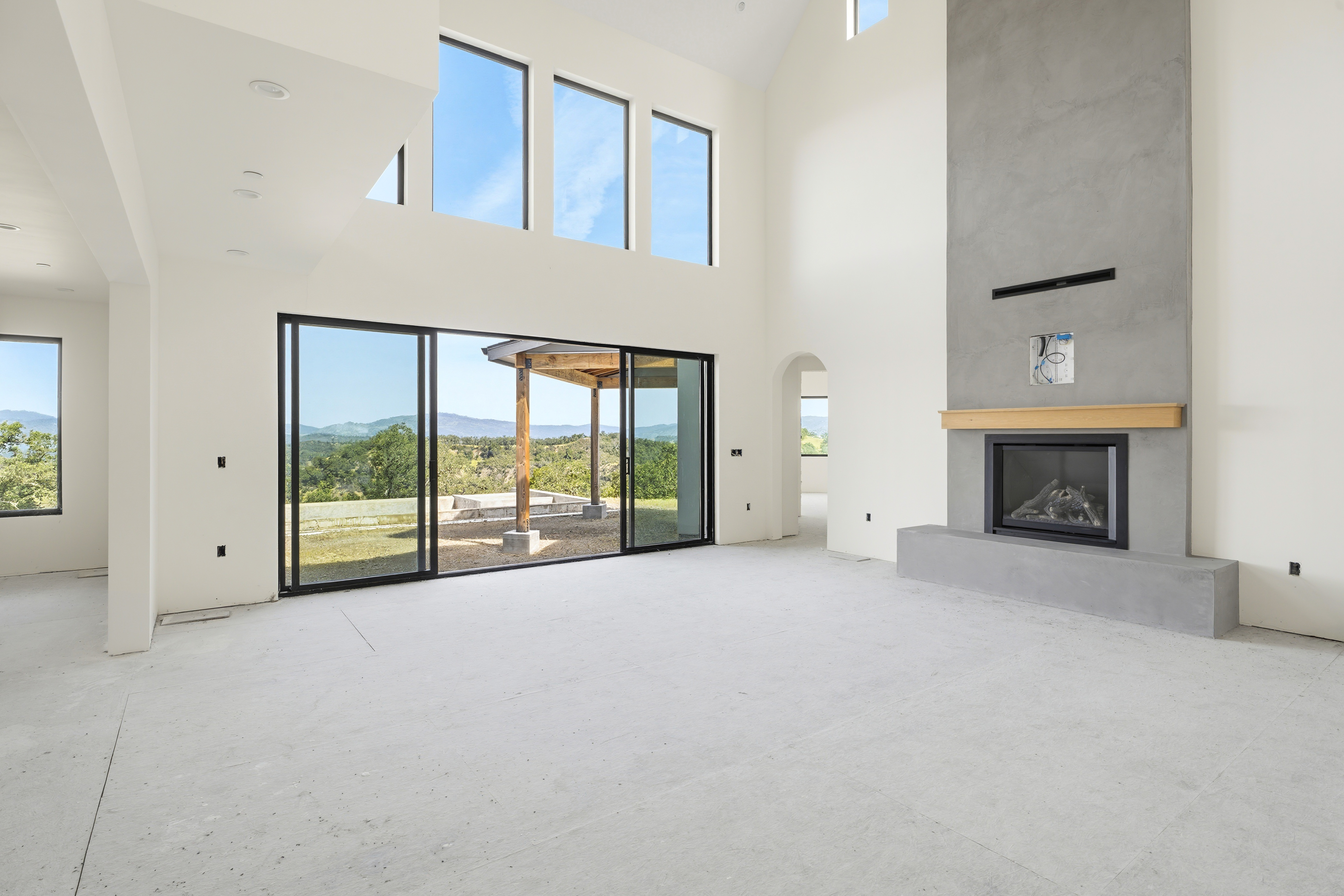 This spacious, high-ceilinged living room features a minimalist aesthetic with unfinished concrete flooring and crisp white walls. A striking floor-to-ceiling gray concrete fireplace serves as the focal point, complemented by expansive sliding glass doors that frame a scenic, elevated view of the surrounding hills. The room is bathed in natural light, emphasizing its airy, open-concept design and potential for modern luxury living.
