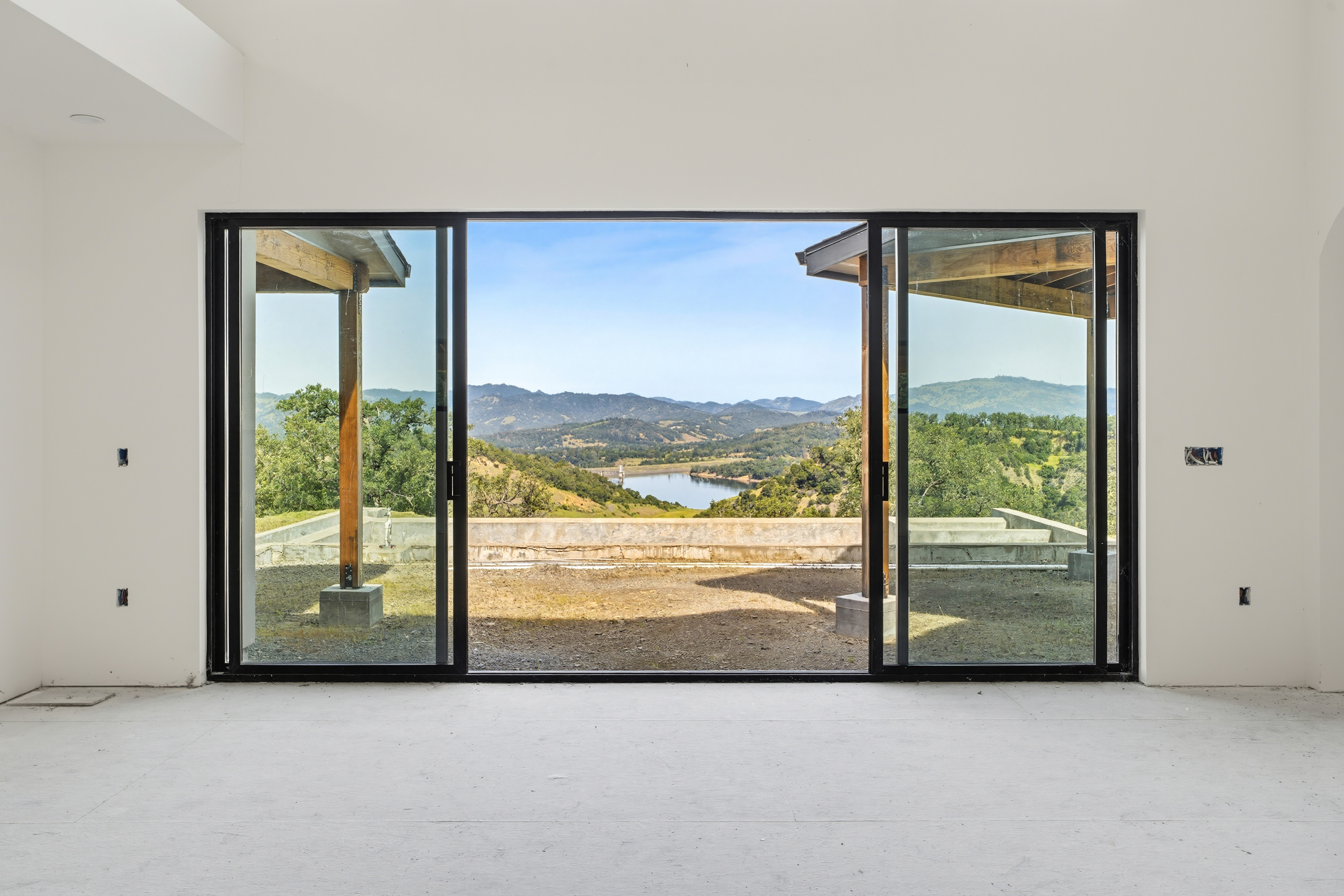 This image captures a minimalist, unfinished interior space featuring a large, black-framed sliding glass door that serves as a dramatic focal point. The expansive opening frames a breathtaking panoramic view of rolling hills, a serene lake, and a partially constructed outdoor patio area. The stark white walls and neutral flooring emphasize the architectural connection between the indoor living space and the natural landscape beyond.