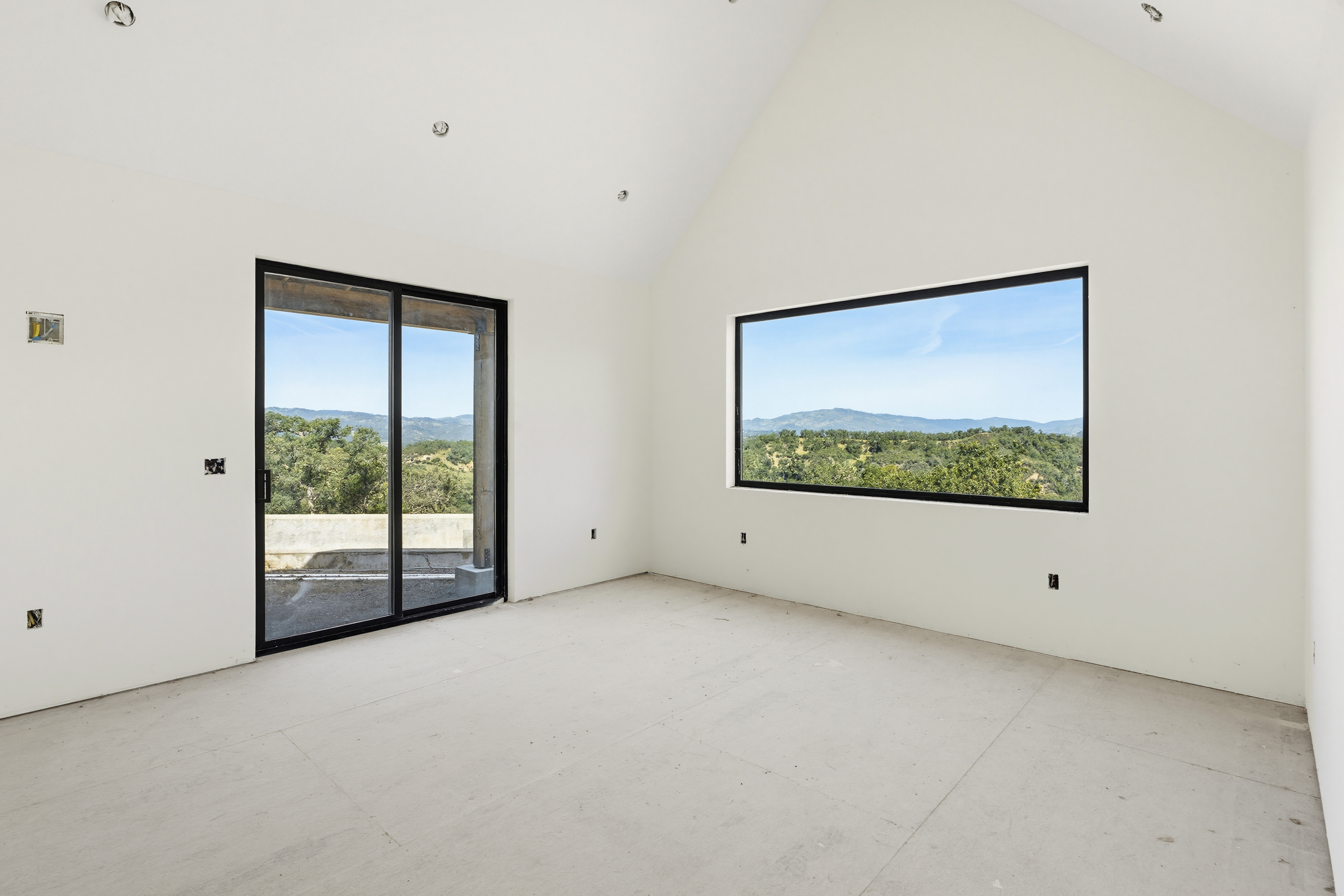 This spacious, unfinished room features a high vaulted ceiling and minimalist white walls, creating a bright and airy atmosphere. A large sliding glass door and a wide rectangular window provide expansive, panoramic views of the surrounding hills and natural landscape. The room is currently in a raw state with unfinished flooring, offering a blank canvas for potential buyers to customize.