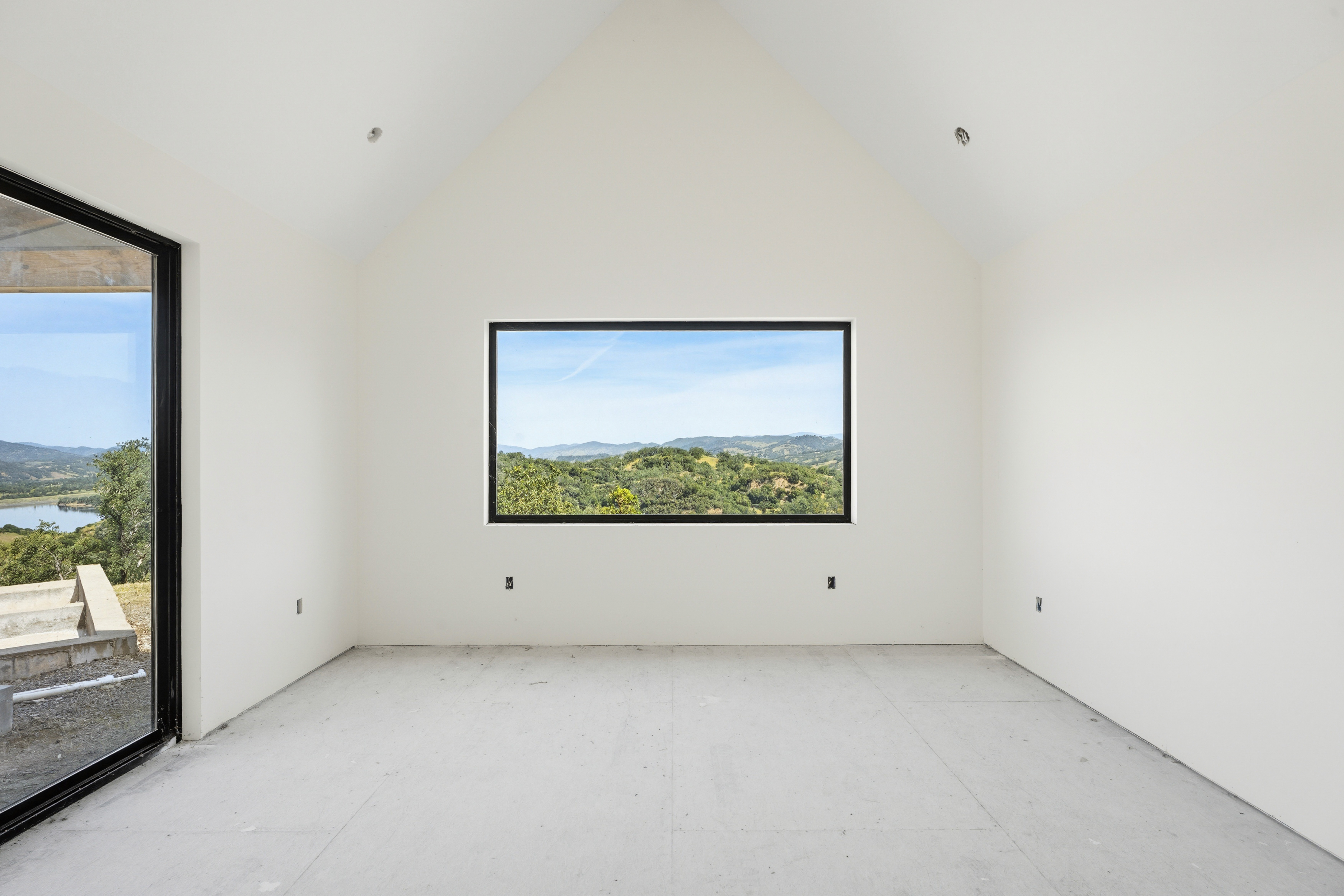This minimalist, unfinished room features a striking vaulted ceiling and a large, rectangular picture window that frames a breathtaking panoramic view of rolling hills and a distant lake. The space is characterized by clean white walls and a neutral concrete floor, offering a blank canvas for a modern interior design. The perspective is centered on the window, emphasizing the seamless connection between the interior architecture and the natural landscape outside.