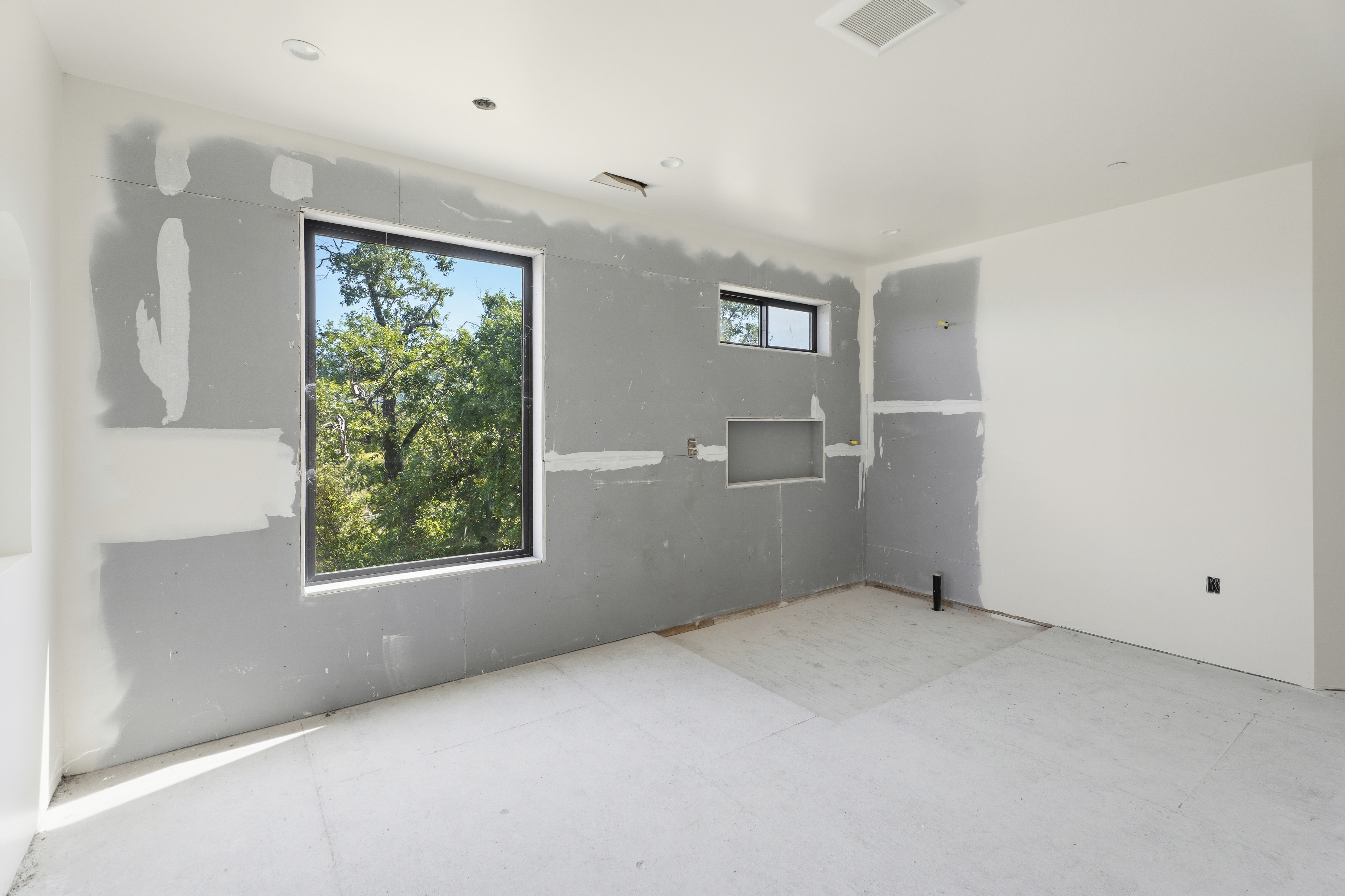 This image shows a primary bedroom under construction, featuring unfinished drywall with visible seams and a large window overlooking a lush, wooded landscape. The room includes a recessed wall niche and a smaller horizontal window, suggesting a modern design layout awaiting final finishes. The perspective is a wide-angle shot capturing the raw, spacious potential of the room.