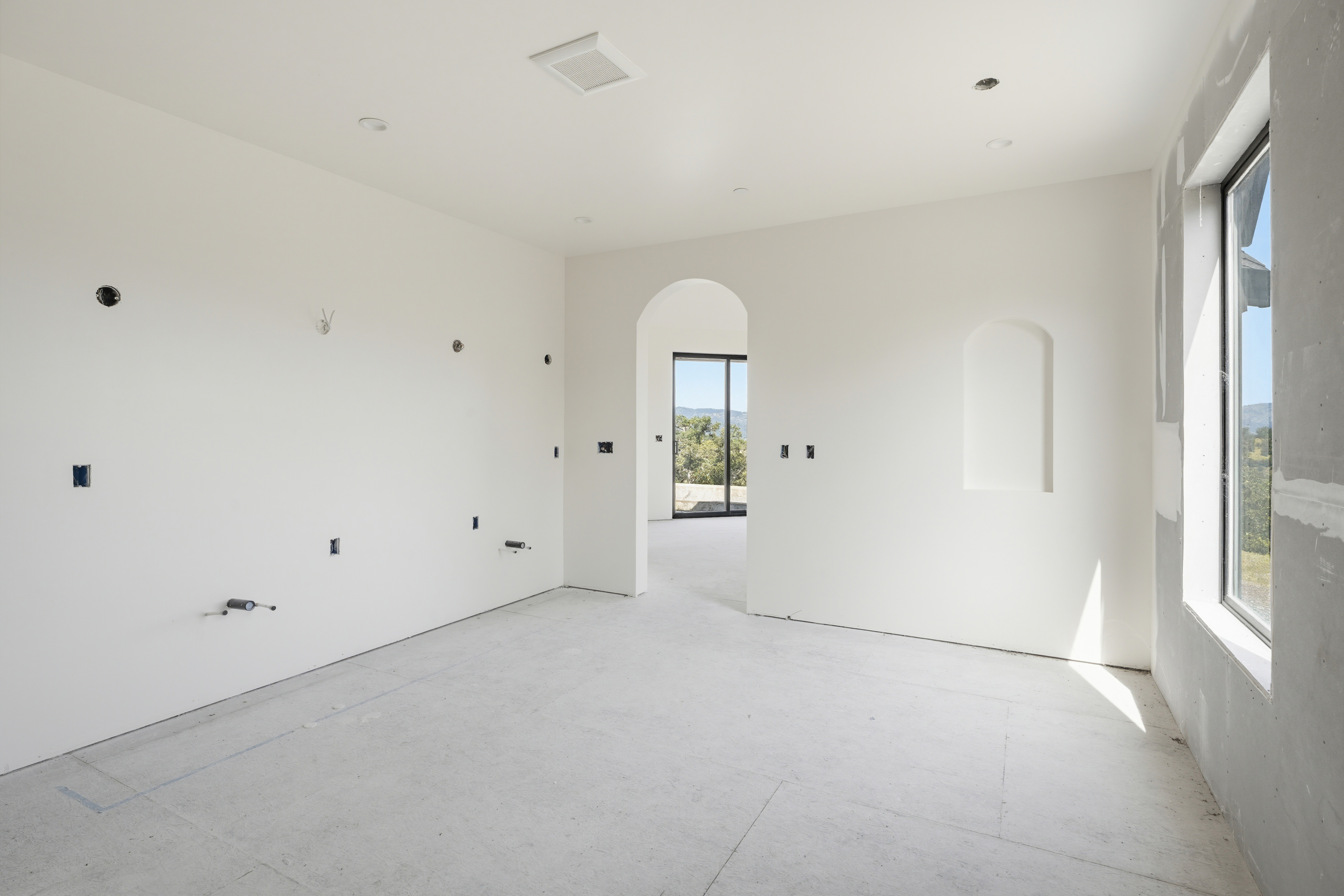 This image shows an unfinished, minimalist kitchen space under construction, featuring white walls and exposed electrical outlets. An arched doorway leads to an adjacent room with a large window, while a recessed wall niche adds architectural interest. The room is bright and airy, highlighting the potential for a modern, custom-designed culinary space.
