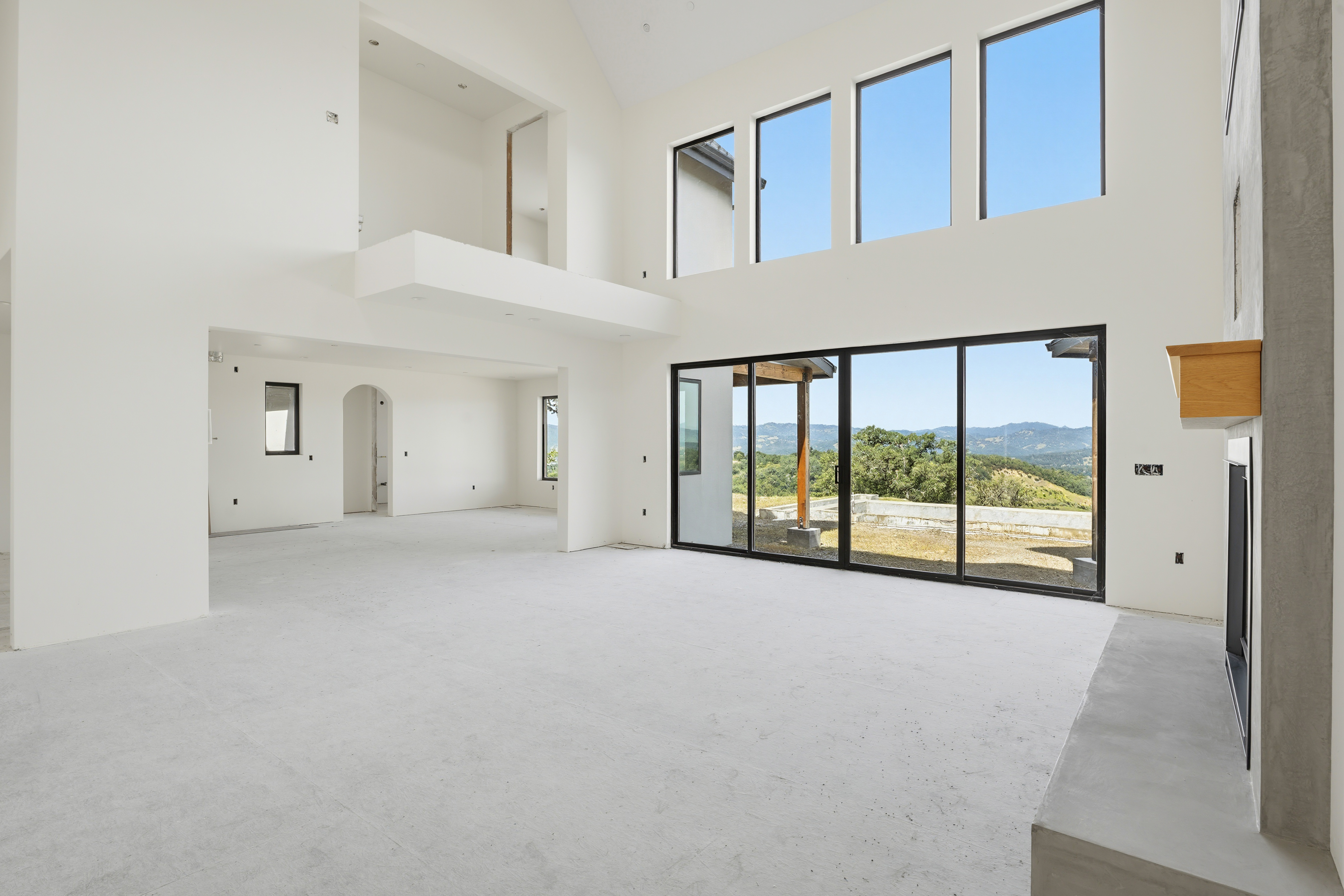 This expansive, high-ceilinged living room features a minimalist, modern aesthetic with white walls and unfinished concrete flooring. A massive wall of floor-to-ceiling windows and sliding glass doors frames a stunning panoramic view of rolling hills and natural landscape. The space includes a prominent concrete fireplace hearth with a wooden mantel, creating a dramatic focal point in this bright, airy, and open-concept architectural design.