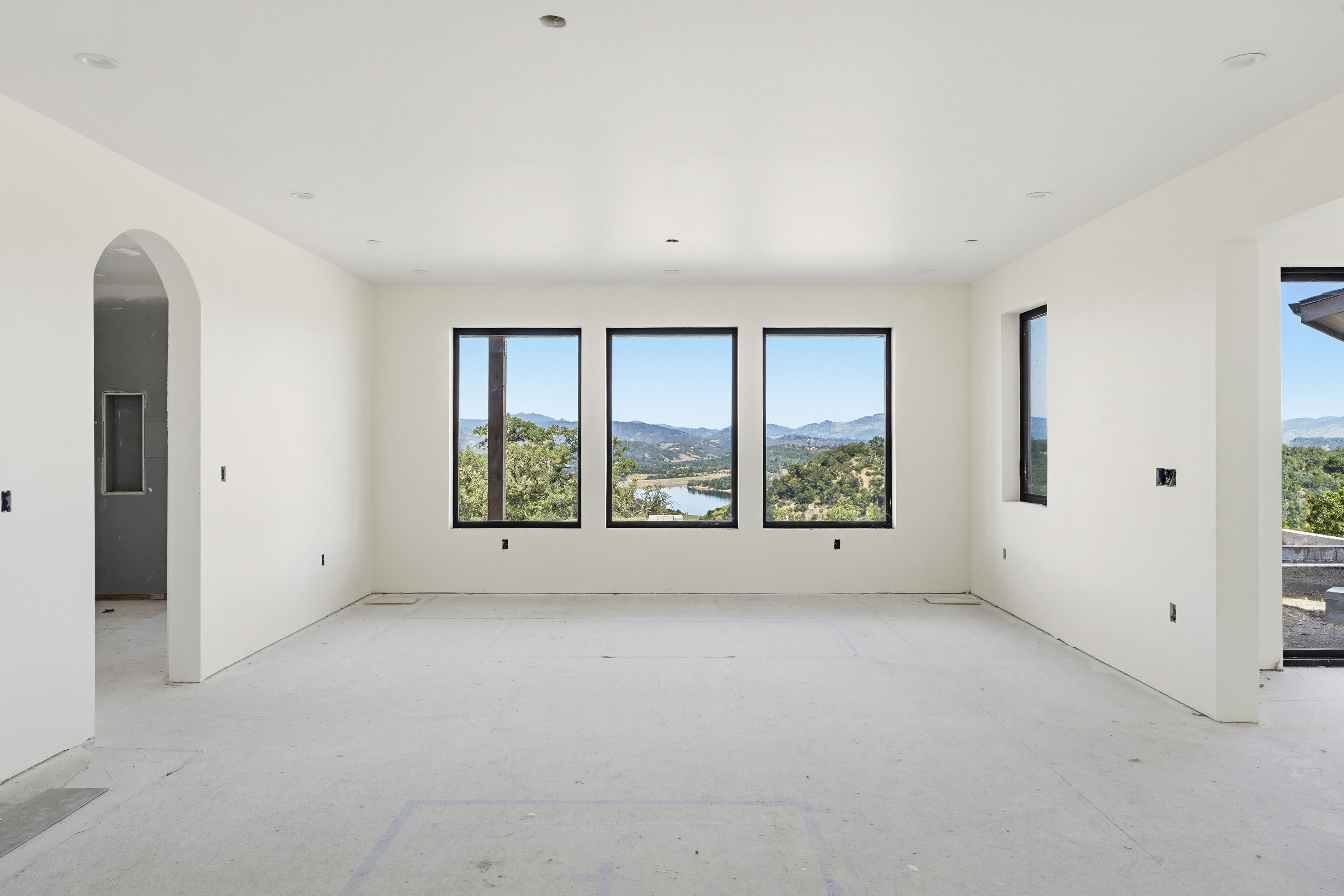 This spacious, unfinished living room features crisp white walls and a neutral concrete floor, providing a blank canvas for customization. Three large, black-framed windows serve as the focal point, framing a stunning panoramic view of rolling hills and a distant lake. An arched doorway to the left adds architectural interest, while the open-concept layout creates an airy and bright atmosphere.