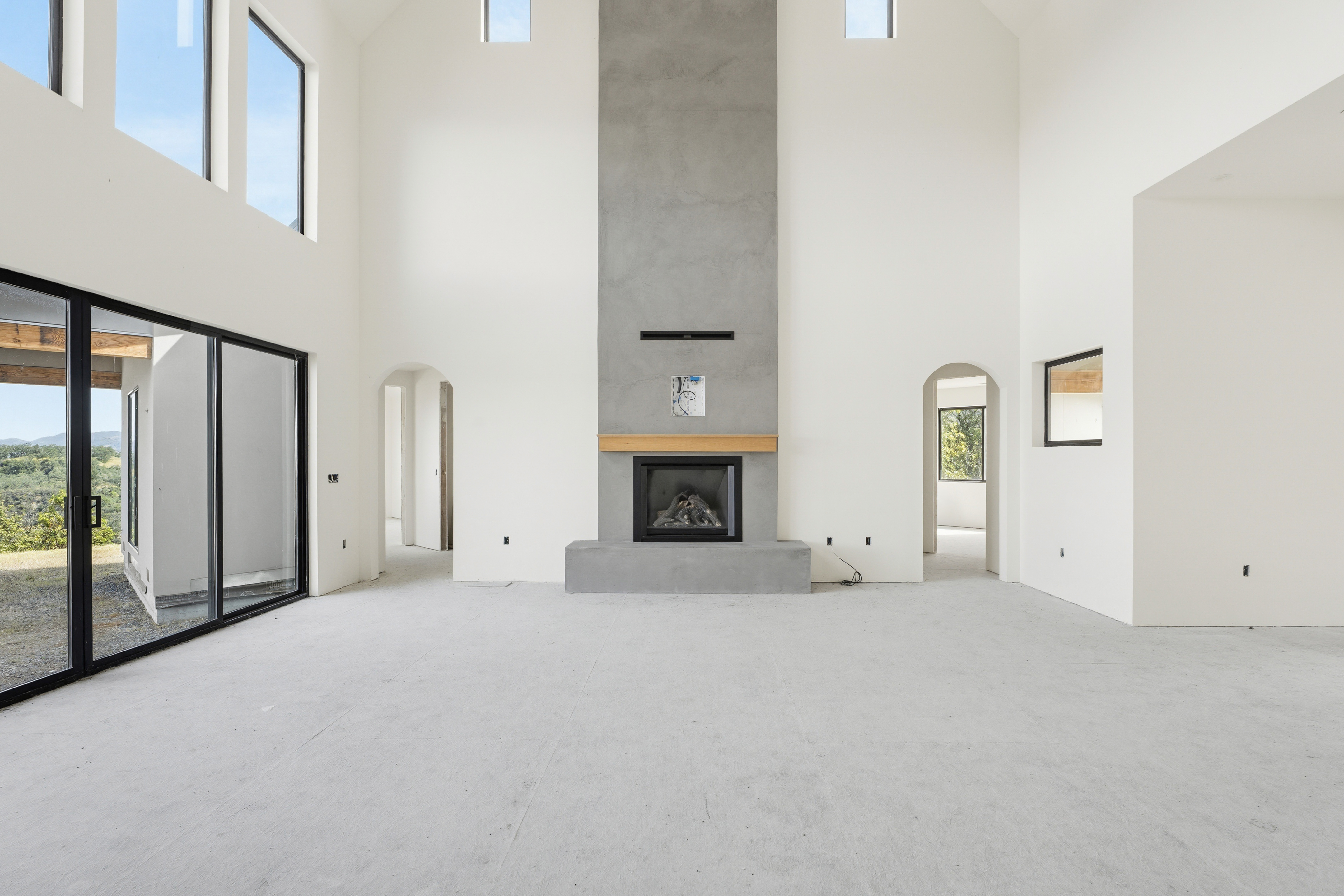 This spacious, high-ceilinged living room features a striking floor-to-ceiling concrete fireplace as its central focal point, accented by a natural wood mantel. The room is bathed in natural light from large, modern windows and glass sliding doors that offer expansive views of the surrounding landscape. With its minimalist white walls, arched doorways, and unfinished concrete flooring, the space presents a clean, contemporary canvas ready for custom interior design.