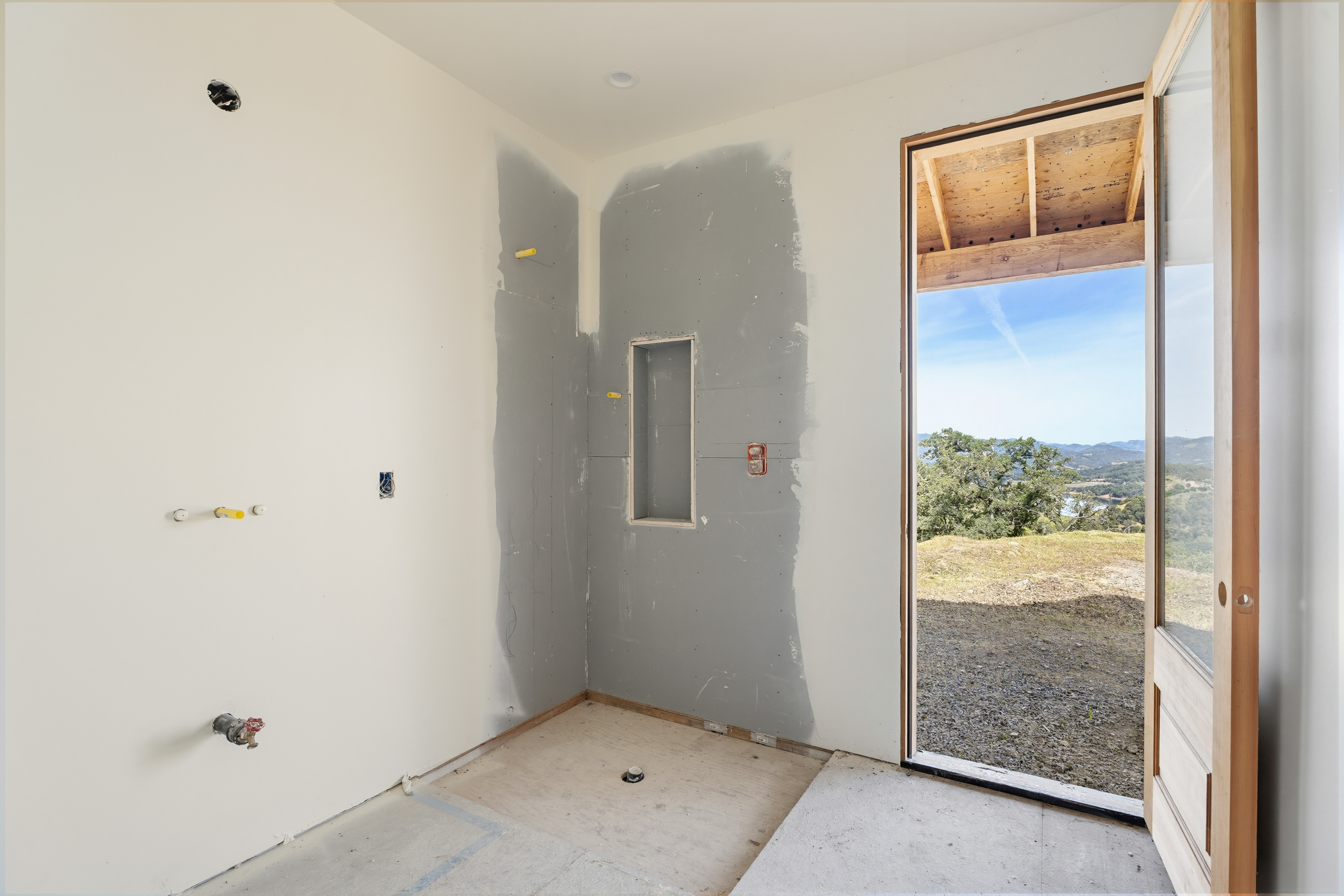 This image captures a bathroom under construction, featuring exposed plumbing, unfinished drywall, and a recessed shower niche. A large, open glass door provides a seamless transition to the outdoors, framing a stunning, expansive view of the surrounding hills and landscape. The space is bright and airy, highlighting the potential for a luxurious, nature-integrated bathroom design.