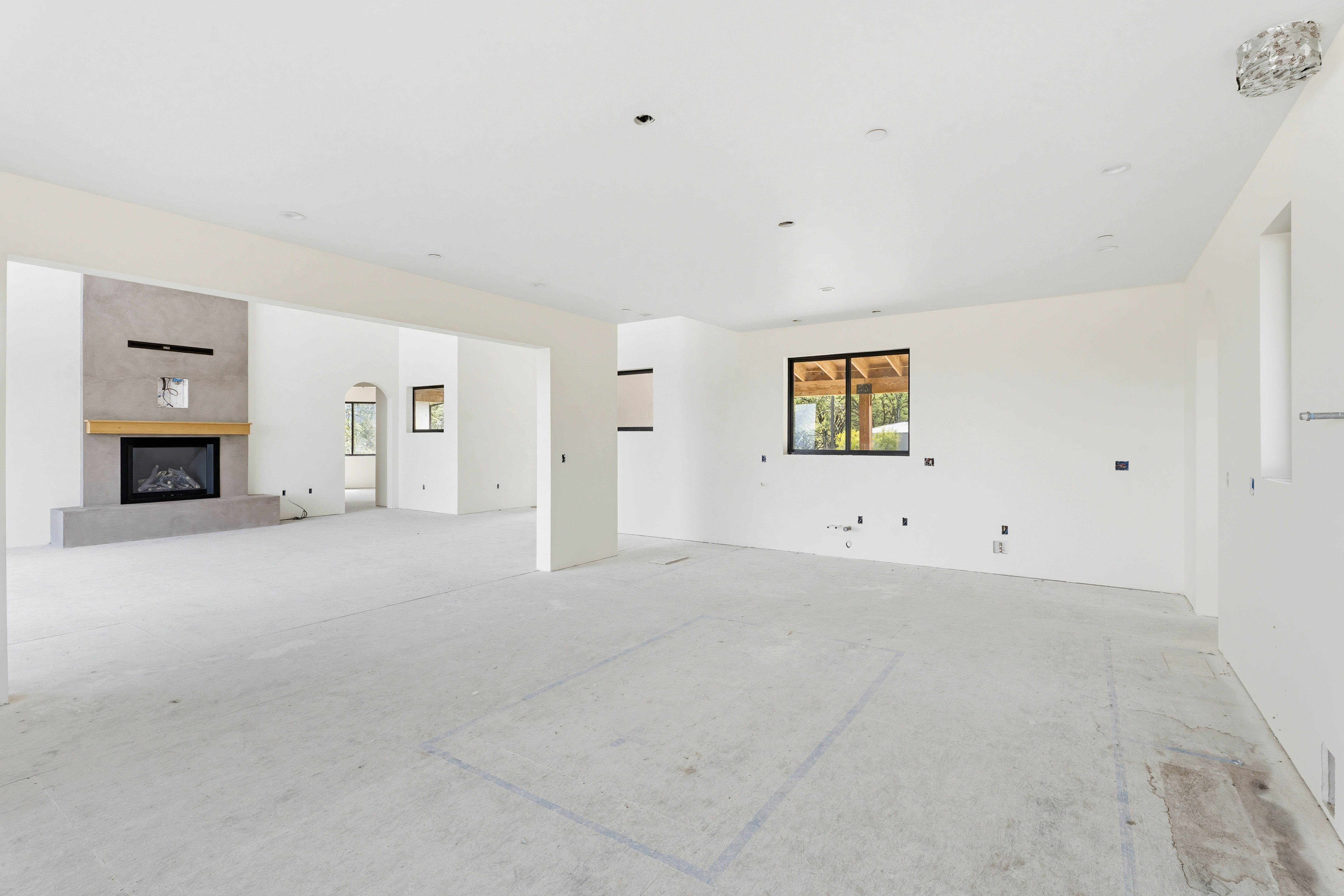 This spacious, open-concept interior is currently under construction, featuring a minimalist aesthetic with white walls and unfinished concrete flooring. A prominent gray fireplace with a wooden mantel serves as a focal point in the adjacent room, while the main area shows layout markings on the floor for future cabinetry or furniture placement. The perspective captures the expansive, airy feel of the home, highlighting its potential for a modern, light-filled living space.