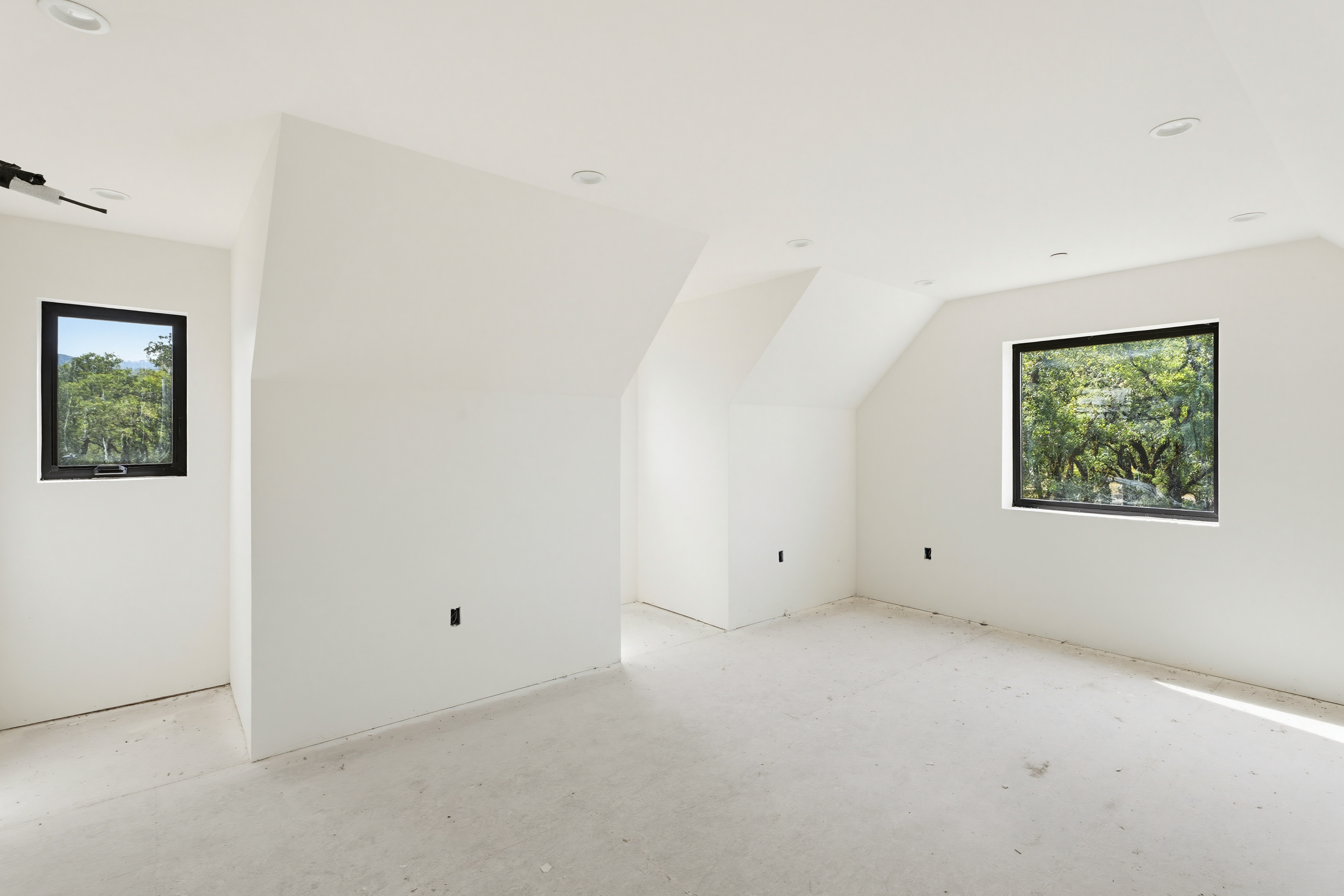 This image showcases an unfinished, minimalist bedroom space characterized by clean white walls and sloped architectural ceilings. Two black-framed windows provide natural light and views of the surrounding greenery, while the neutral, unfinished flooring suggests a blank canvas ready for customization. The perspective is wide and centered, emphasizing the room's unique geometry and potential for a modern living area.