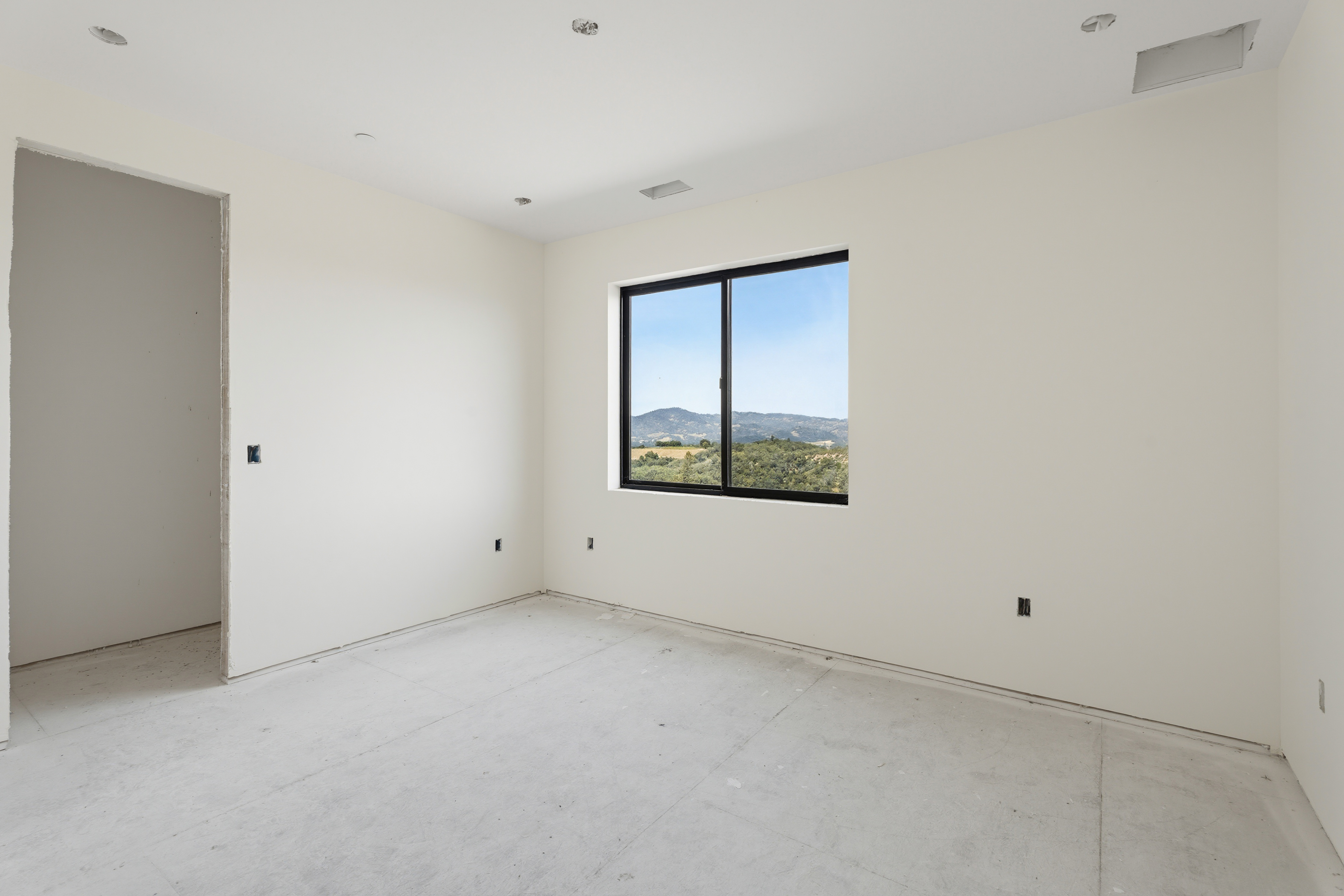 This image shows an unfinished bedroom interior featuring plain, off-white walls and a concrete subfloor. A single, modern window with a black frame serves as the focal point, offering a clear view of the surrounding hills and landscape. The room is currently in a construction phase, highlighting the potential for customization in a new build.