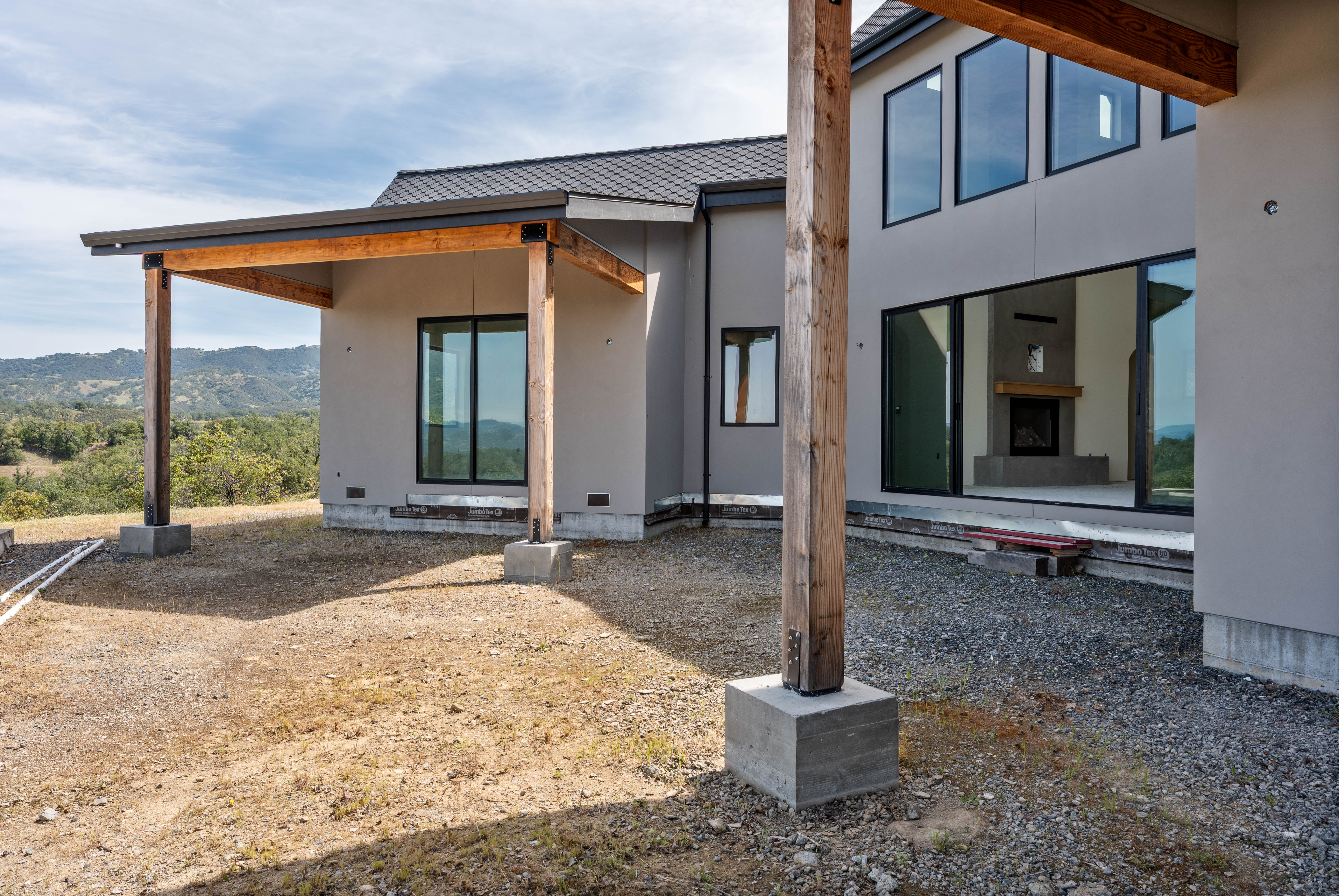 This rear view of a modern, contemporary home showcases a spacious covered patio area supported by rustic timber beams and concrete footings. The exterior features smooth, light-toned stucco walls, large black-framed windows, and a sleek dark roof, with an unfinished gravel yard extending toward a scenic, rolling hillside landscape. The perspective captures the transition between the indoor living space, visible through the large sliding glass doors, and the expansive natural surroundings.