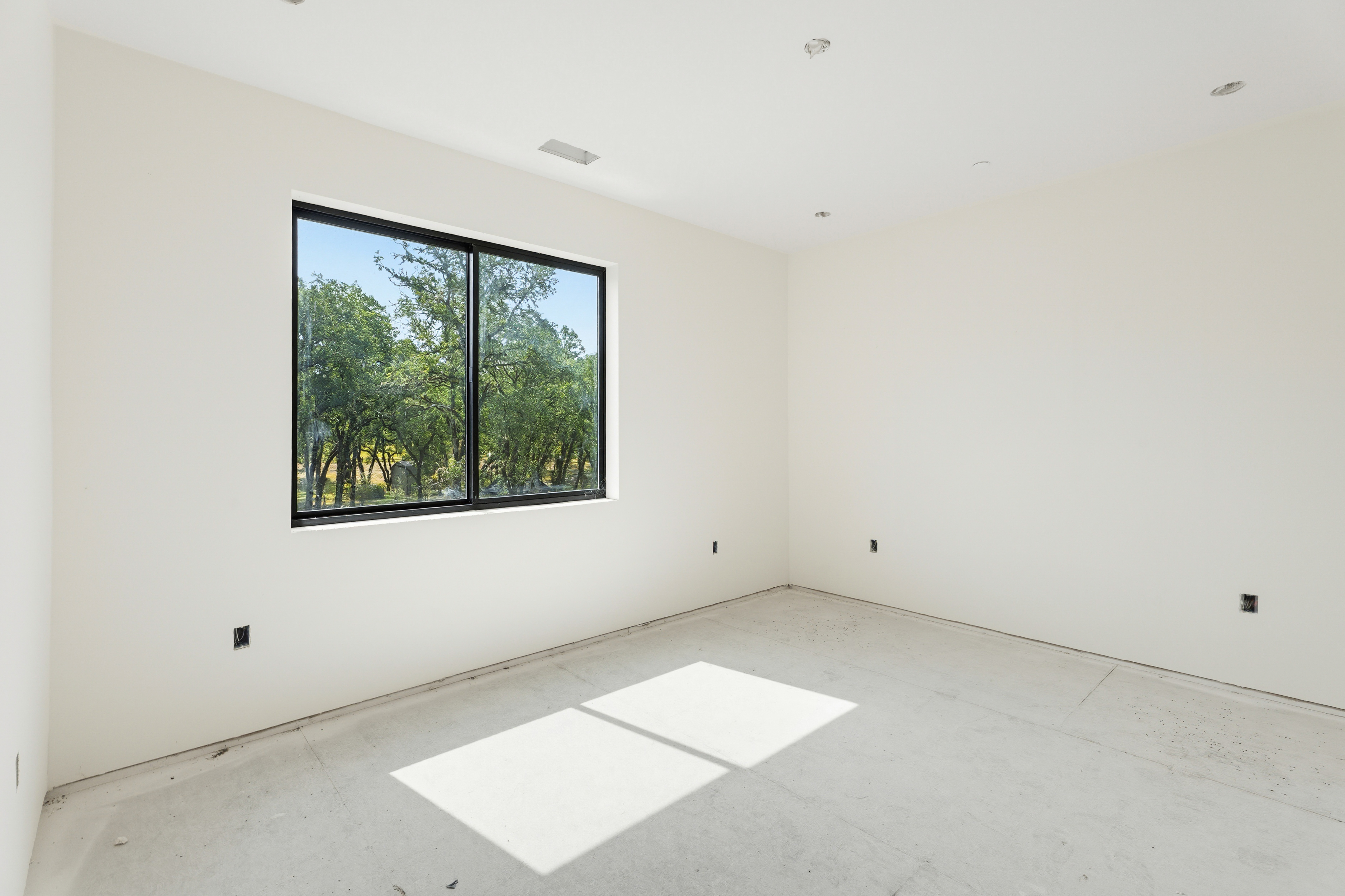 This image captures a bright, unfinished bedroom space featuring clean, white walls and a concrete subfloor. A large, black-framed window serves as the focal point, offering a serene view of lush green trees outside and casting a distinct rectangular patch of sunlight onto the floor. The room is a blank canvas, highlighting the potential for customization in a modern, minimalist home.