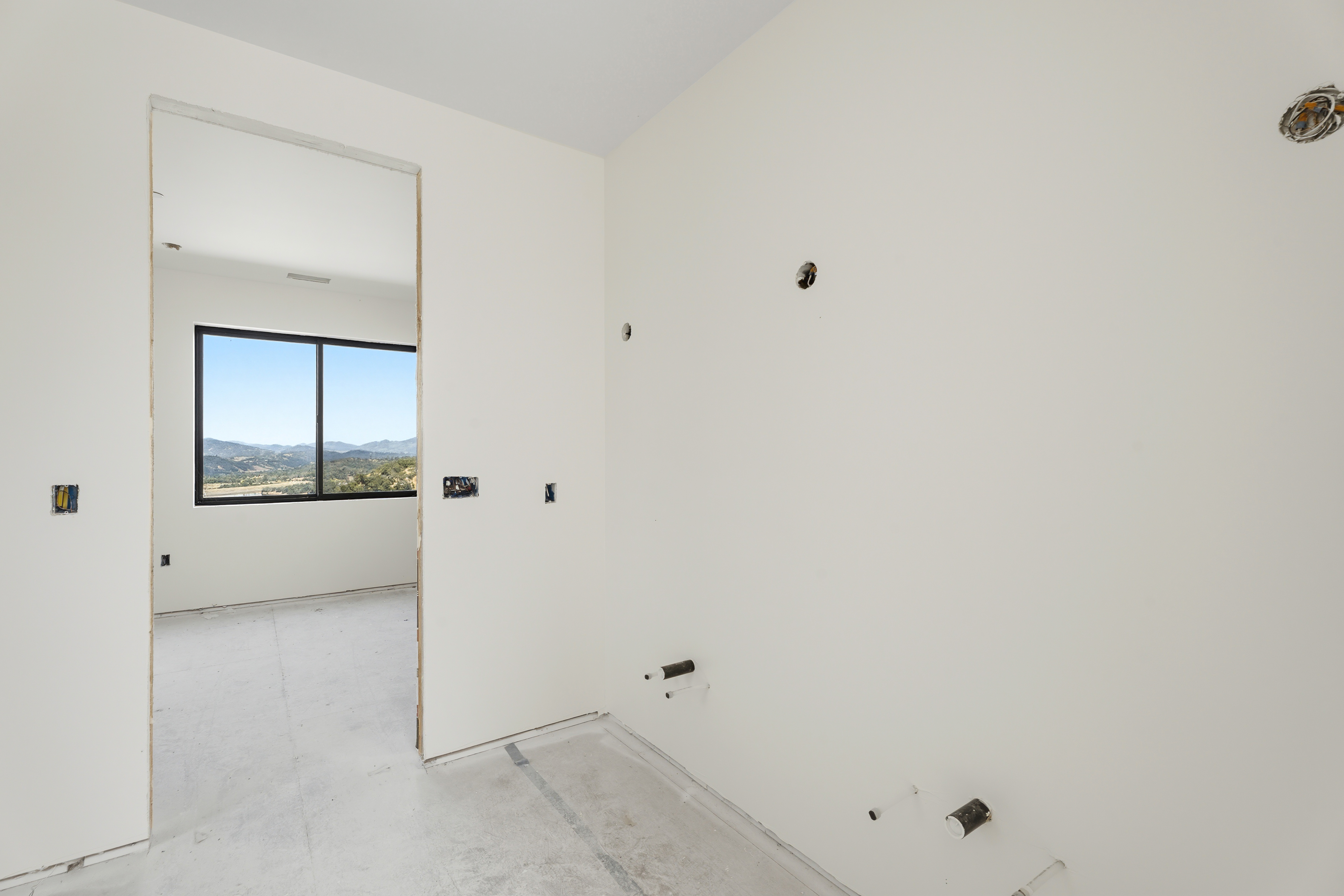 This image shows an unfinished bathroom space under construction, featuring exposed plumbing pipes and electrical wiring on plain white walls. A doorway leads into an adjacent room with a large window that provides a scenic view of distant hills and landscape. The room is currently in a raw, preparatory state, highlighting the potential for a custom design.