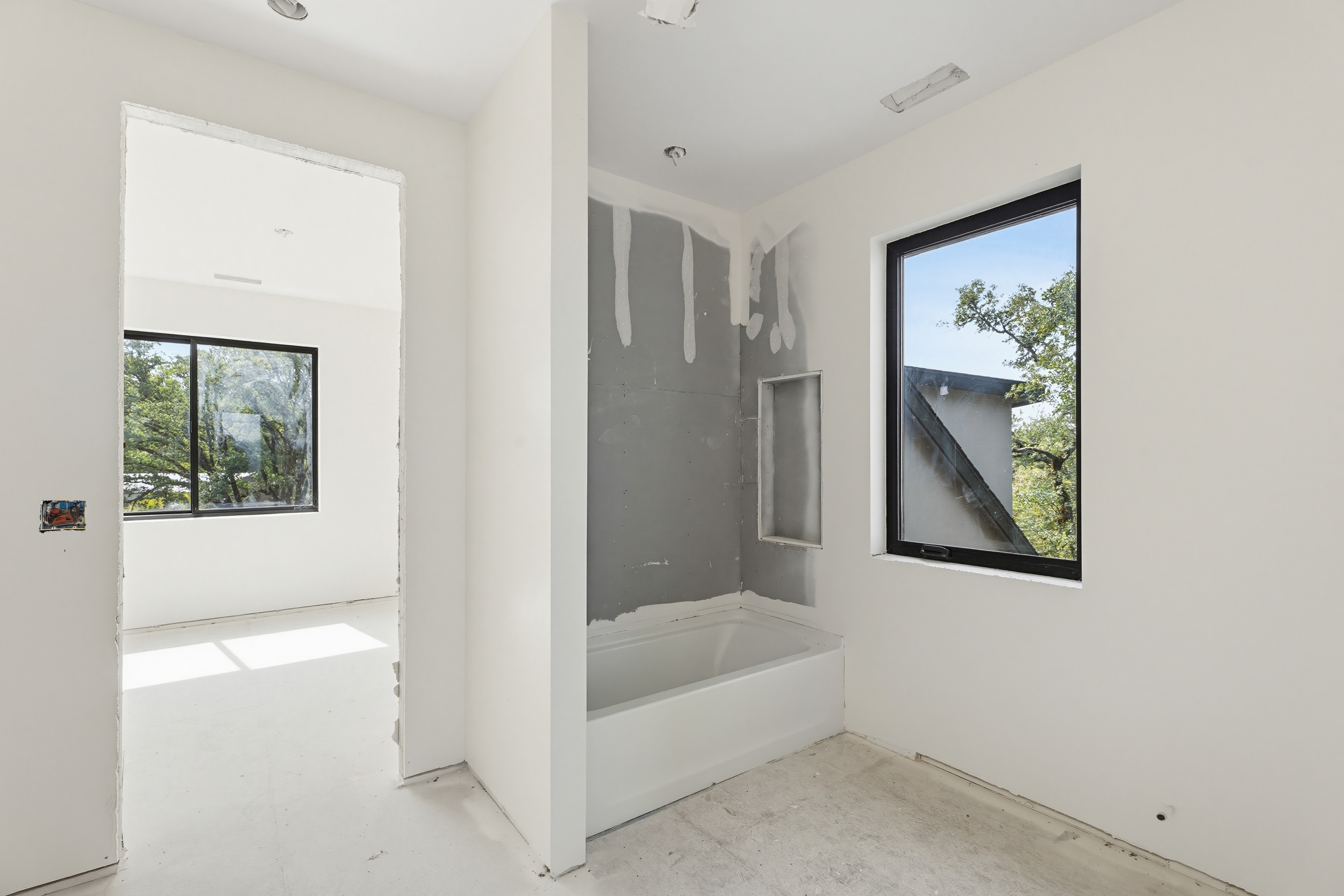 This image captures a bathroom in the midst of construction, featuring a white bathtub installed against a grey, unfinished wall with a built-in shower niche. The space is bright and minimalist, with white walls and a large window that provides a view of the surrounding trees. The perspective is from the doorway, looking into the bathroom and through to an adjacent room, highlighting the open, airy layout of the home.