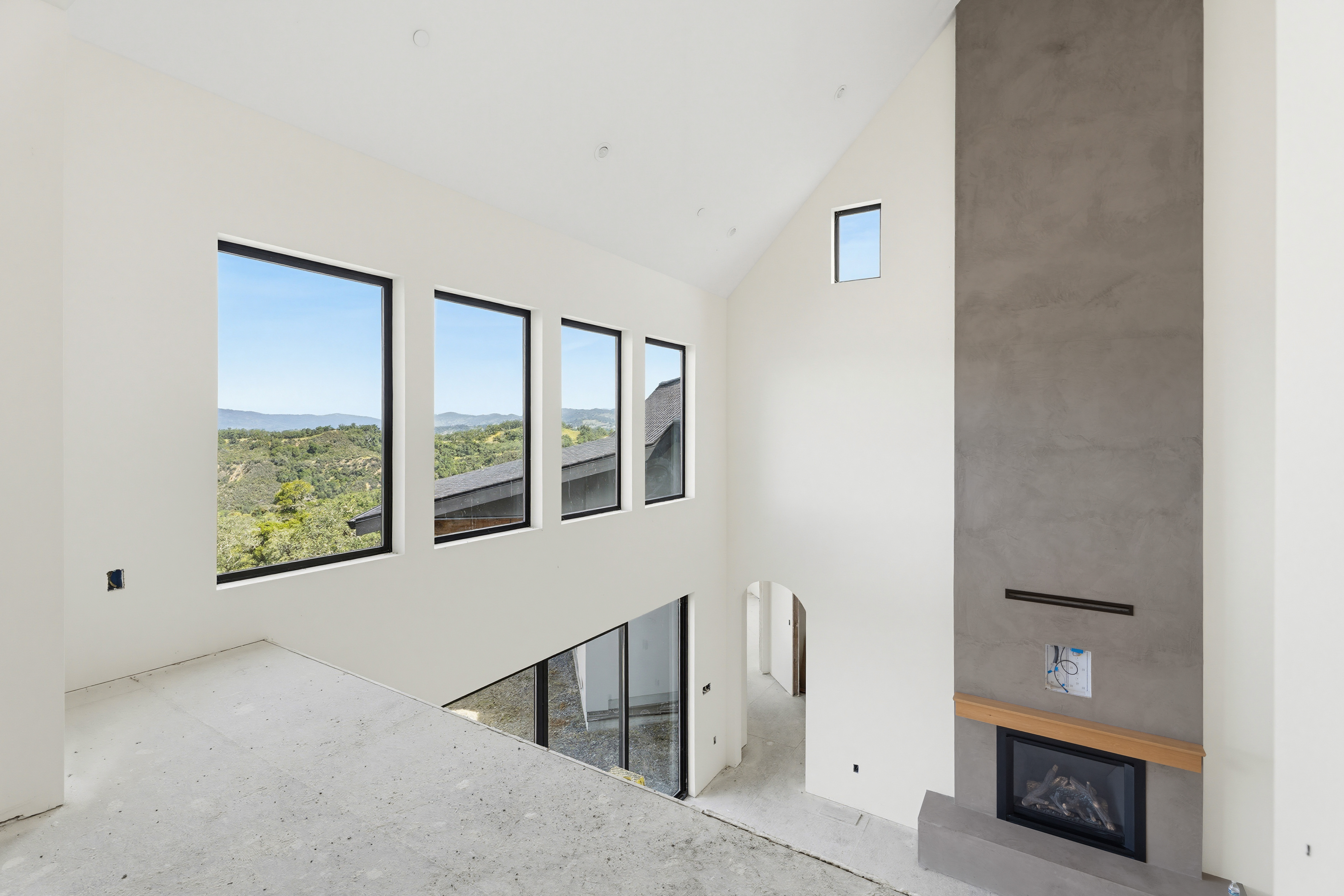 This high-ceilinged, modern living room is currently under construction, featuring a striking floor-to-ceiling concrete fireplace as its focal point. Large, expansive windows flood the space with natural light and offer panoramic views of the surrounding hills, while the open-concept design connects the upper level to the lower floor. The minimalist aesthetic and raw materials create a sophisticated, airy atmosphere that highlights the home's architectural potential.