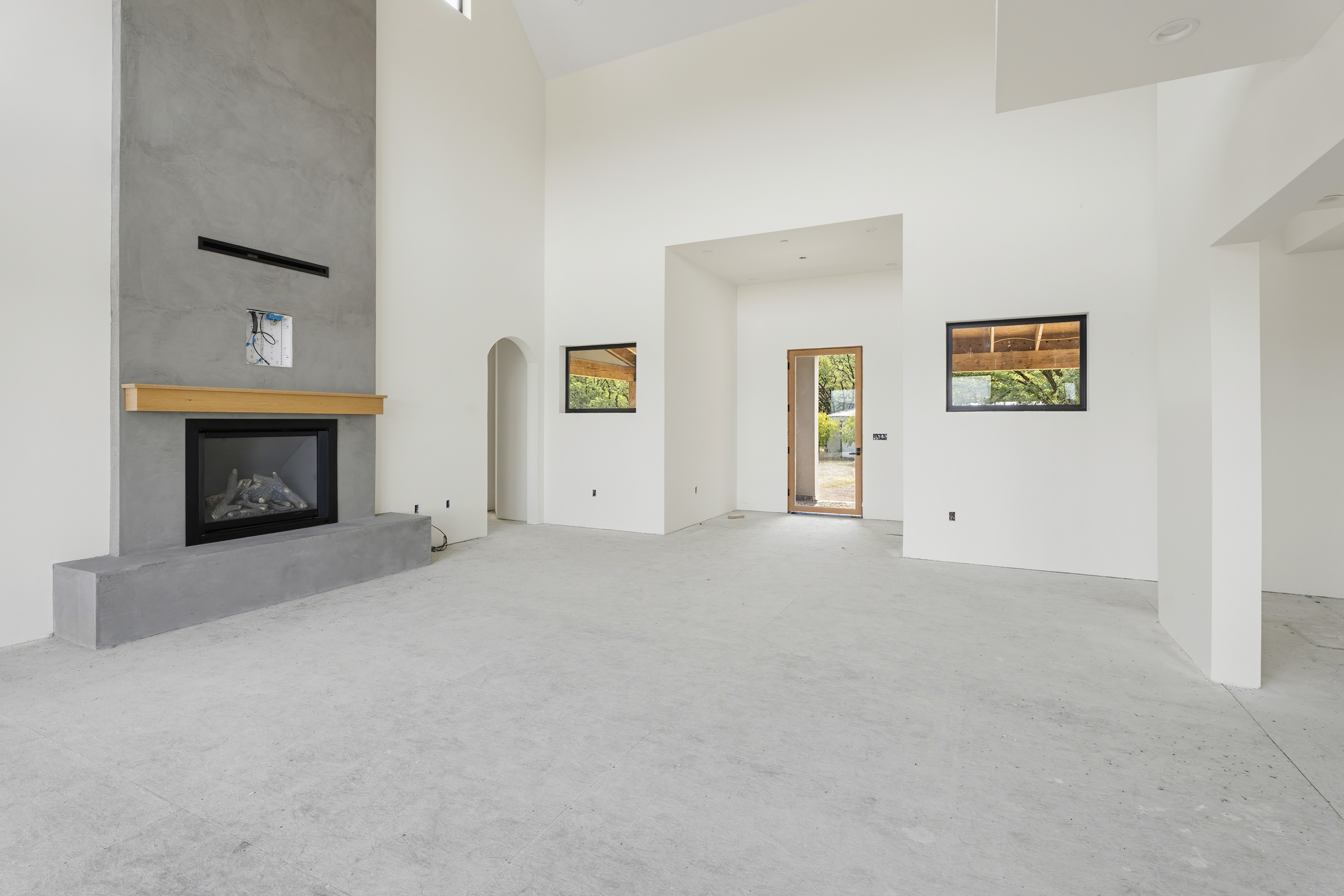 This spacious, high-ceilinged living room features a striking floor-to-ceiling concrete fireplace as its focal point, complemented by a minimalist aesthetic. The room is bathed in natural light from multiple windows and a glass-paneled door, highlighting the clean white walls and unfinished concrete flooring. The open-concept layout and architectural details, such as the arched doorway, suggest a modern, airy design ready for custom interior styling.