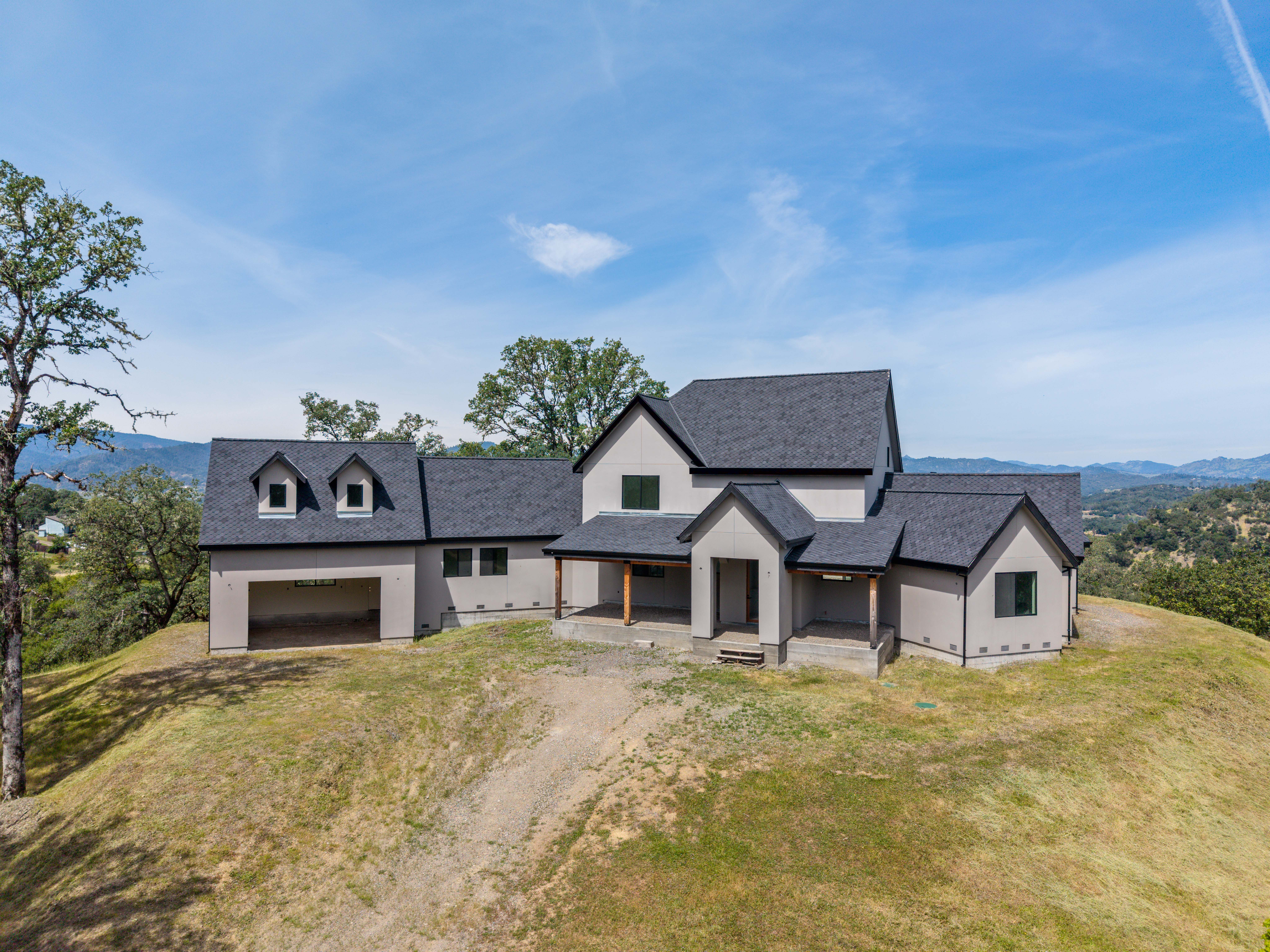 This aerial perspective captures a modern, newly constructed home perched atop a grassy hill, showcasing its contemporary farmhouse aesthetic with dark shingled roofing and neutral-toned stucco siding. The structure features a prominent garage, a covered front porch with wooden support beams, and multiple gabled rooflines that create a dynamic silhouette against the expansive blue sky. The surrounding landscape is open and hilly, emphasizing the property's secluded and scenic location.