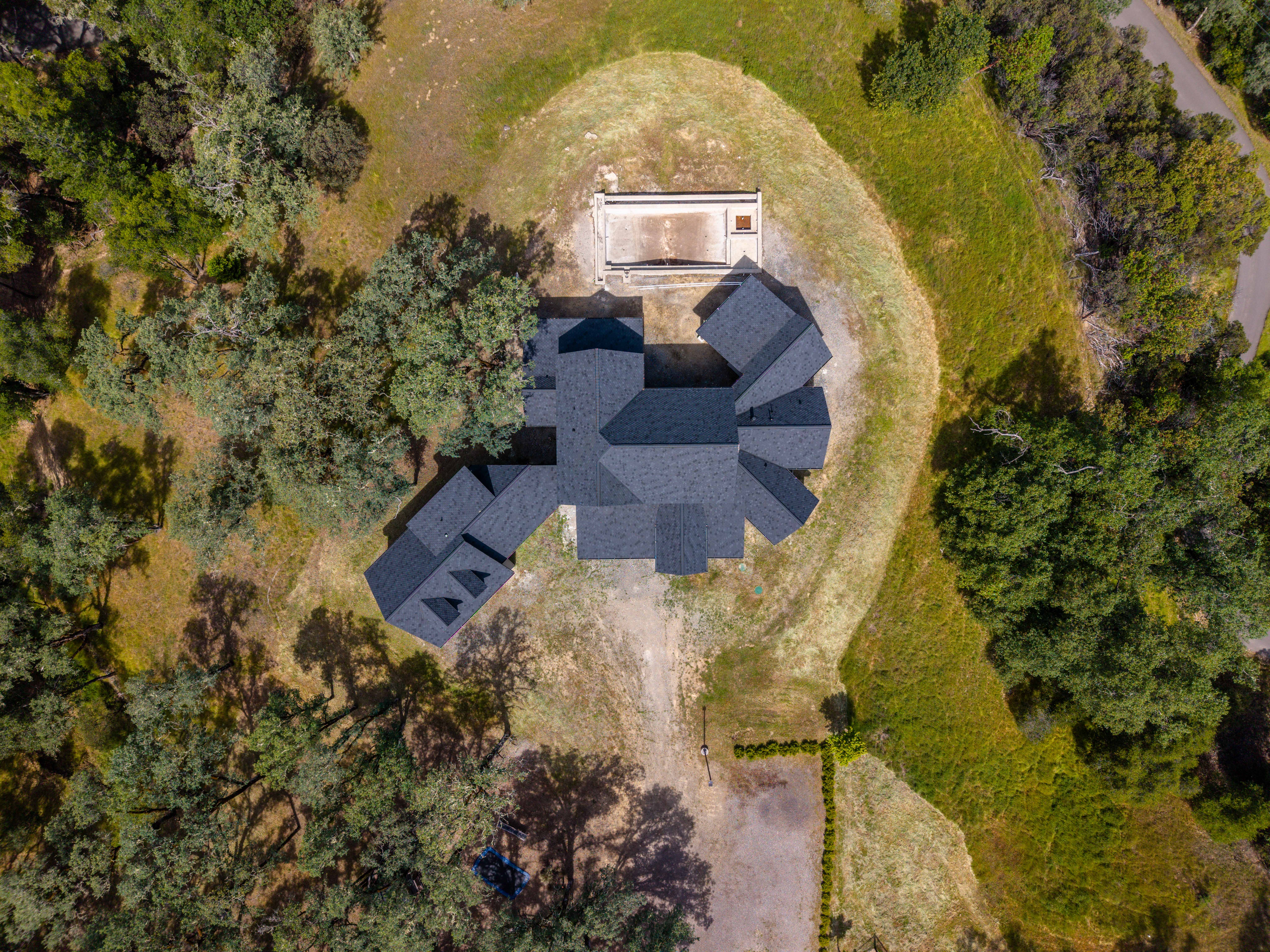 This high-angle aerial perspective captures a sprawling, multi-gabled residential property nestled within a lush, wooded landscape. The home features a complex dark-shingled roofline and is situated on a large, private lot that includes an unfinished concrete pool structure in the rear. The surrounding terrain is characterized by rolling grassy hills and mature oak trees, emphasizing a secluded and serene estate setting.