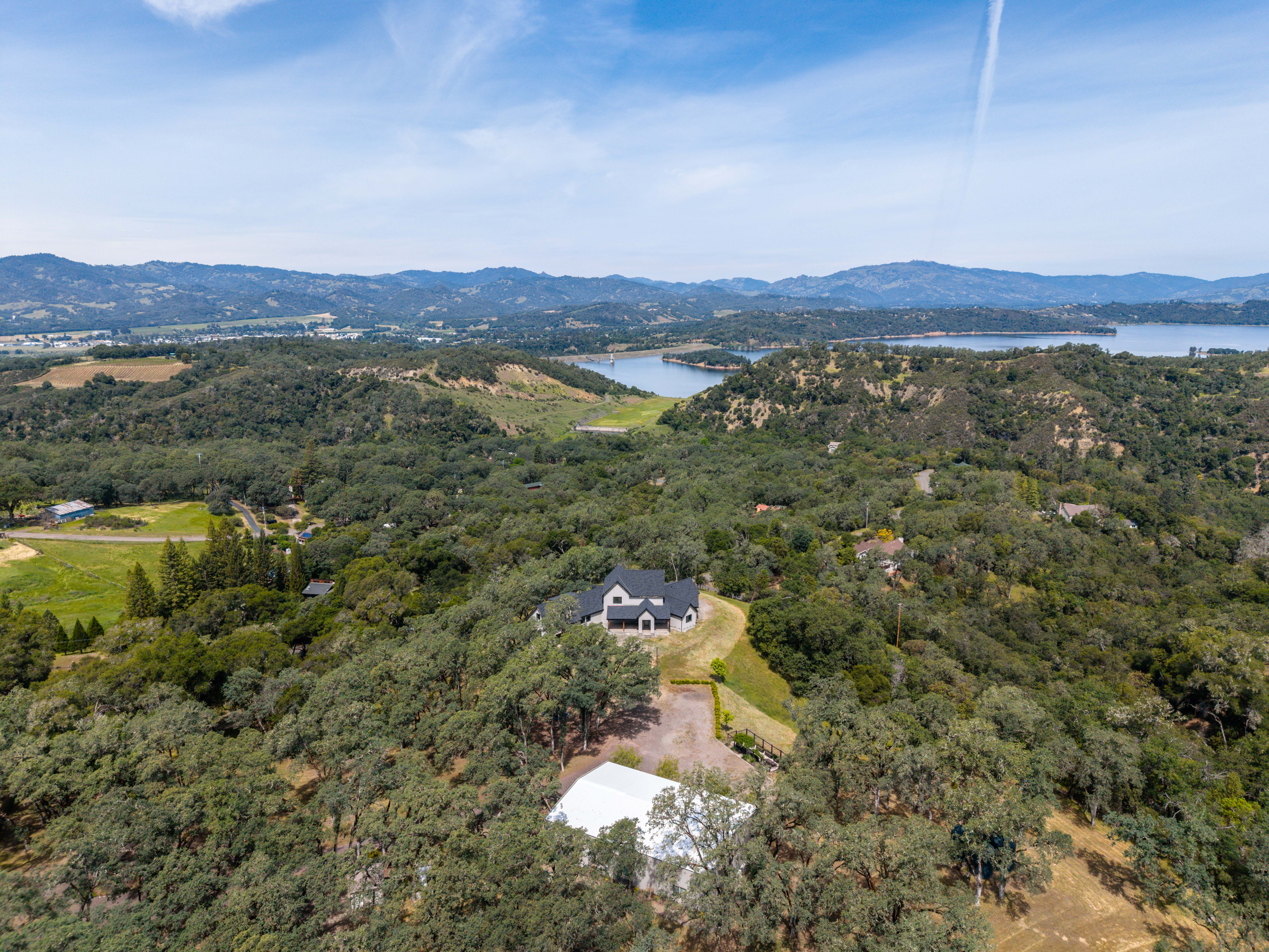 This high-angle aerial shot captures a sprawling rural property nestled within a dense, wooded landscape. The scene features a primary residence with a dark roof and a separate large white outbuilding, both surrounded by mature trees and rolling hills that lead to a distant lake. The perspective emphasizes the expansive, secluded nature of the estate and its picturesque natural surroundings.