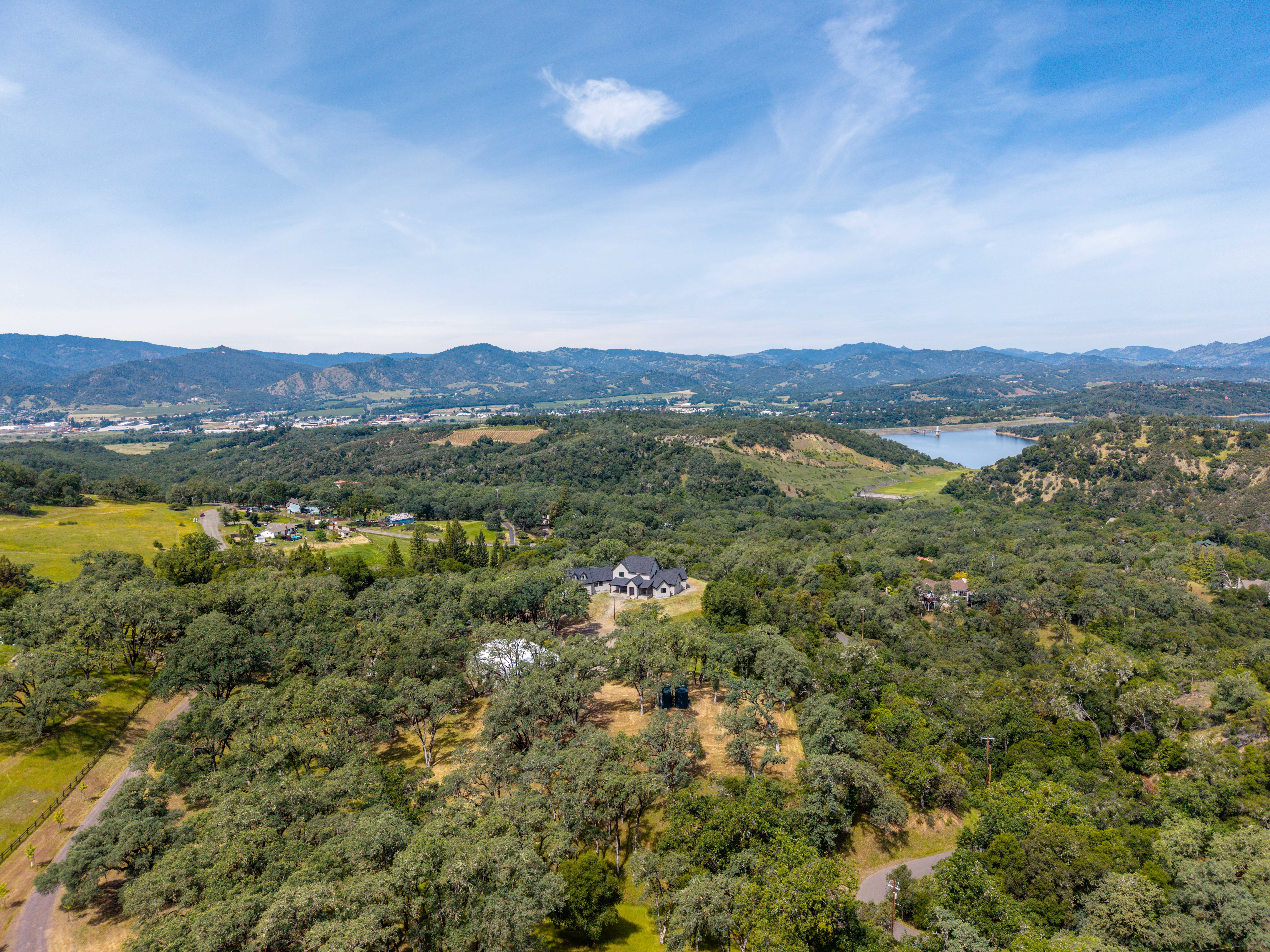 This high-angle aerial shot captures a sprawling rural estate nestled within a lush, tree-covered landscape. The property features a modern farmhouse-style residence surrounded by rolling hills, with a scenic reservoir visible in the distance under a bright, expansive sky. The perspective emphasizes the property's secluded and peaceful setting, highlighting its integration with the natural environment.