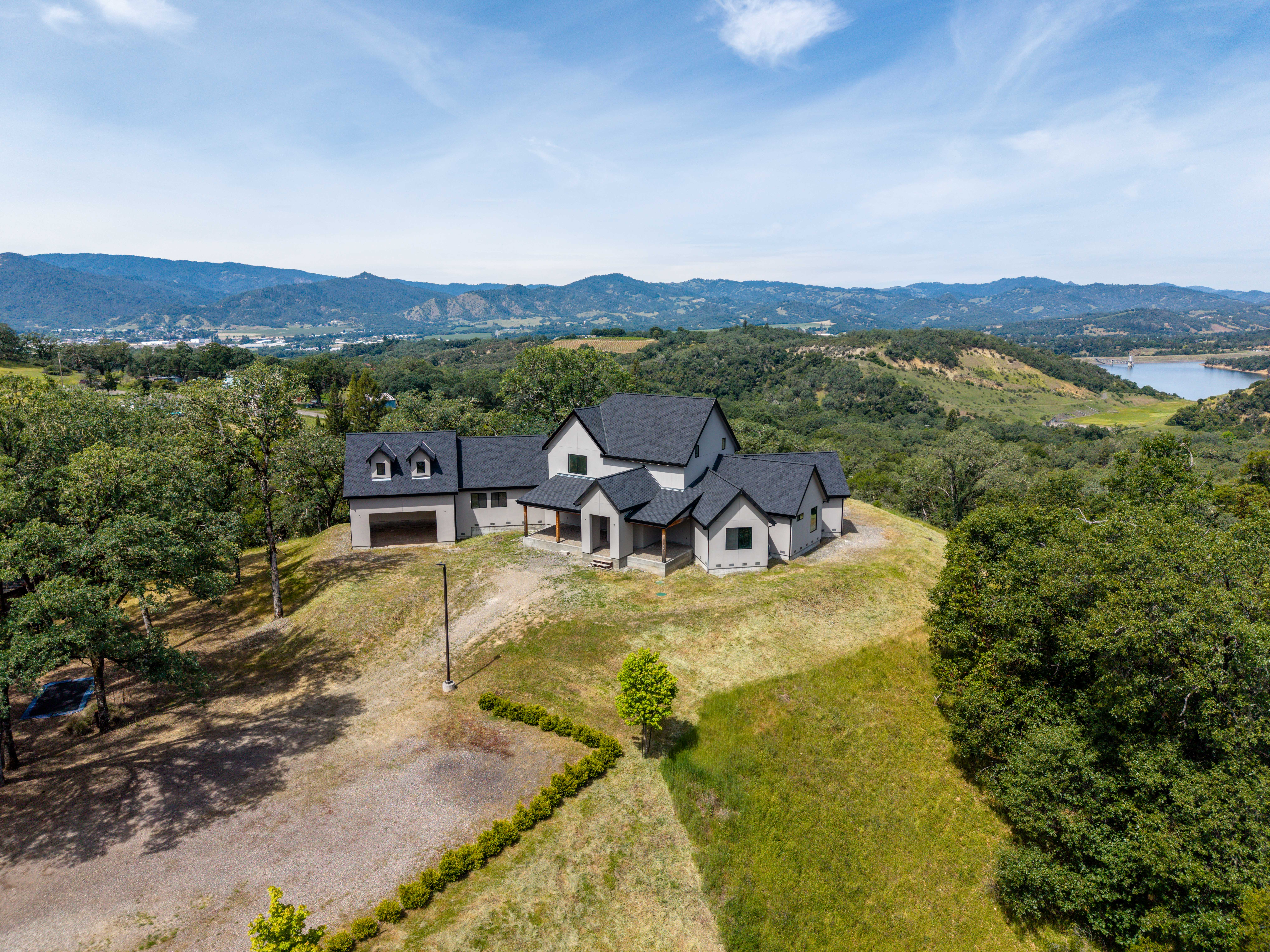 This high-angle aerial shot captures a modern farmhouse-style residence situated prominently on a grassy hilltop. The home features a multi-gabled roofline with dark shingles, light-colored siding, and a detached garage, all set against a sprawling landscape of rolling hills and a distant lake. The perspective emphasizes the property's secluded, scenic location and its impressive architectural scale.