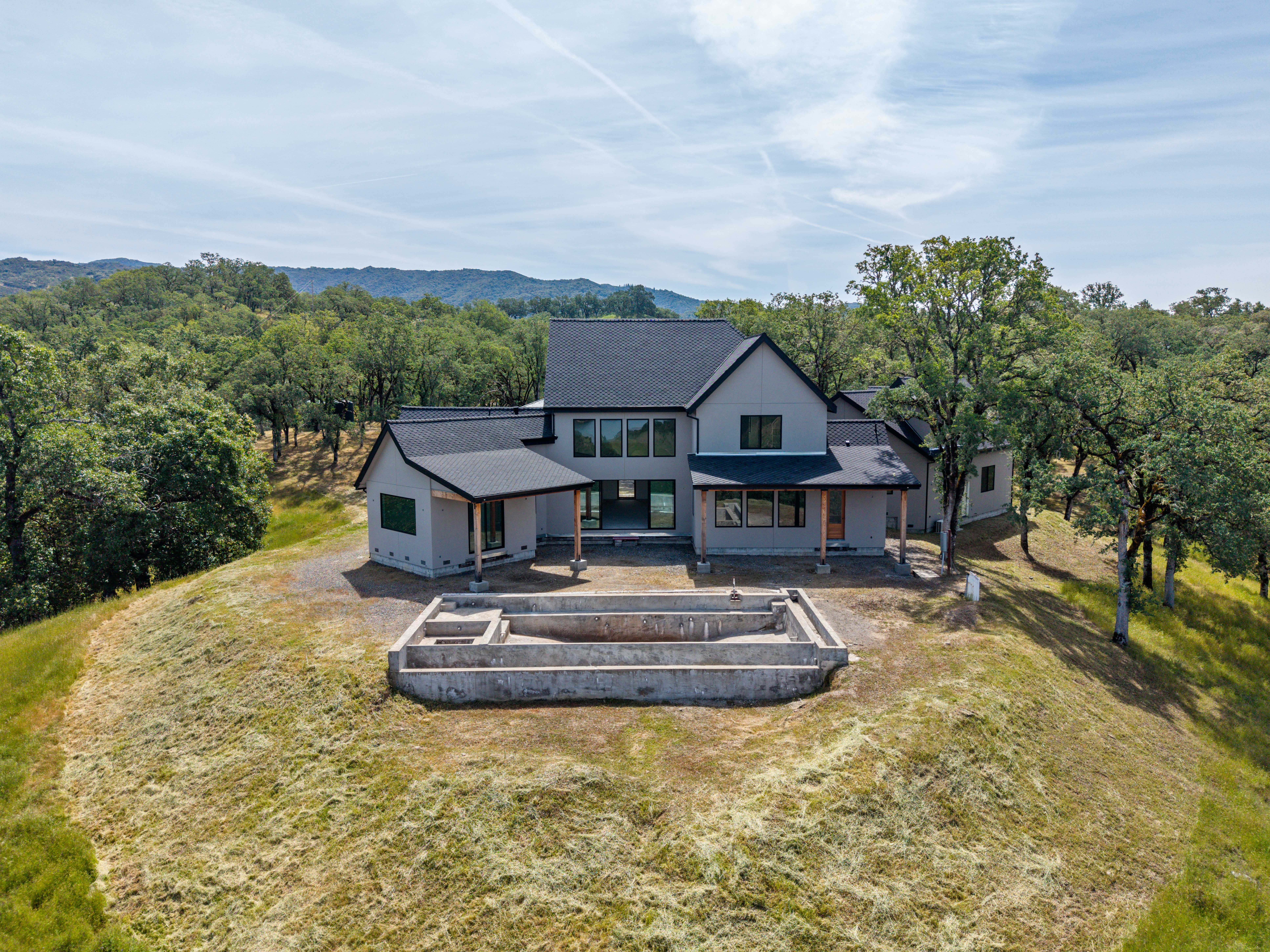 This aerial perspective captures a modern, multi-gabled home under construction, situated prominently on a grassy hilltop surrounded by lush, rolling woodland. In the foreground, a large, unfinished concrete pool structure awaits completion, emphasizing the property's potential for outdoor living. The home features a sleek gray exterior with dark roofing, blending contemporary design with the natural, expansive landscape.
