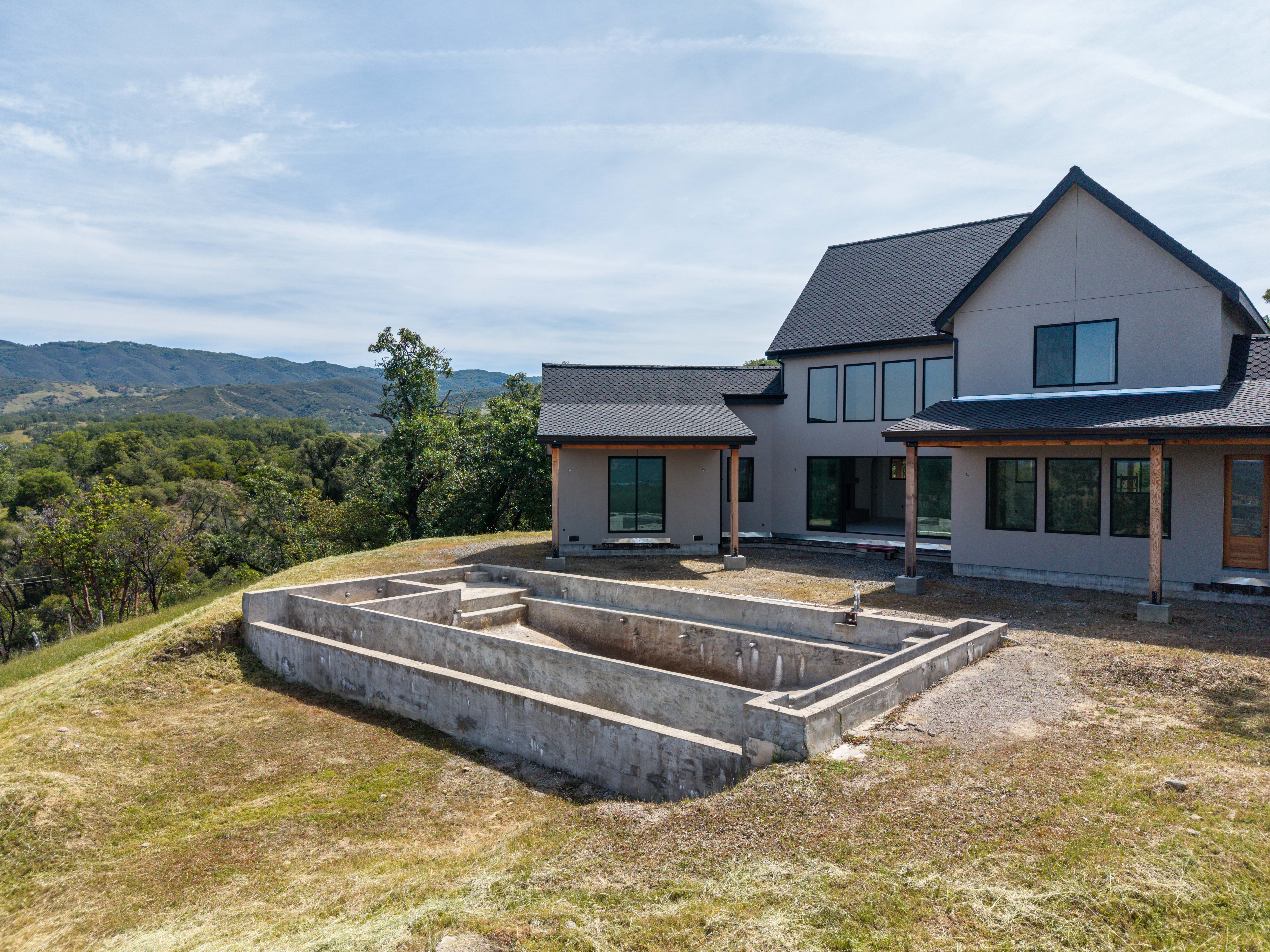 This aerial perspective captures a modern, two-story home under construction, set against a backdrop of rolling hills and lush greenery. In the foreground, a large, unfinished concrete swimming pool structure sits prominently on the hillside, highlighting the potential for a luxury outdoor living space. The home features clean lines, dark roofing, and large windows, suggesting a contemporary architectural style that blends with the natural landscape.