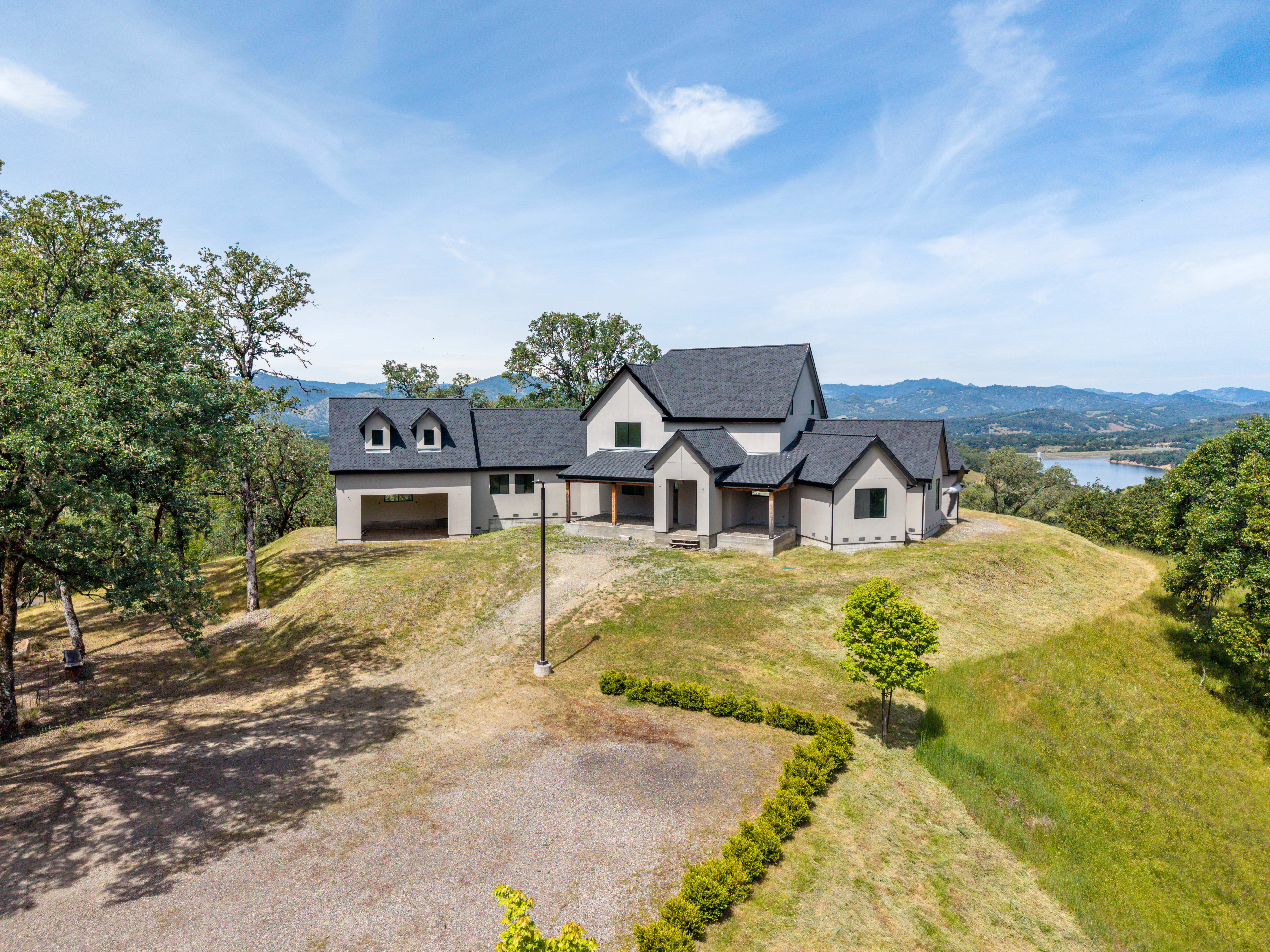 This high-angle aerial shot captures a modern, multi-gabled farmhouse-style home perched prominently on a grassy hilltop. The property features a neutral-toned exterior with dark roofing, a spacious garage, and a covered front porch, all set against a backdrop of rolling hills and a distant lake. The wide-angle perspective emphasizes the home's secluded, scenic location and its integration into the natural landscape.