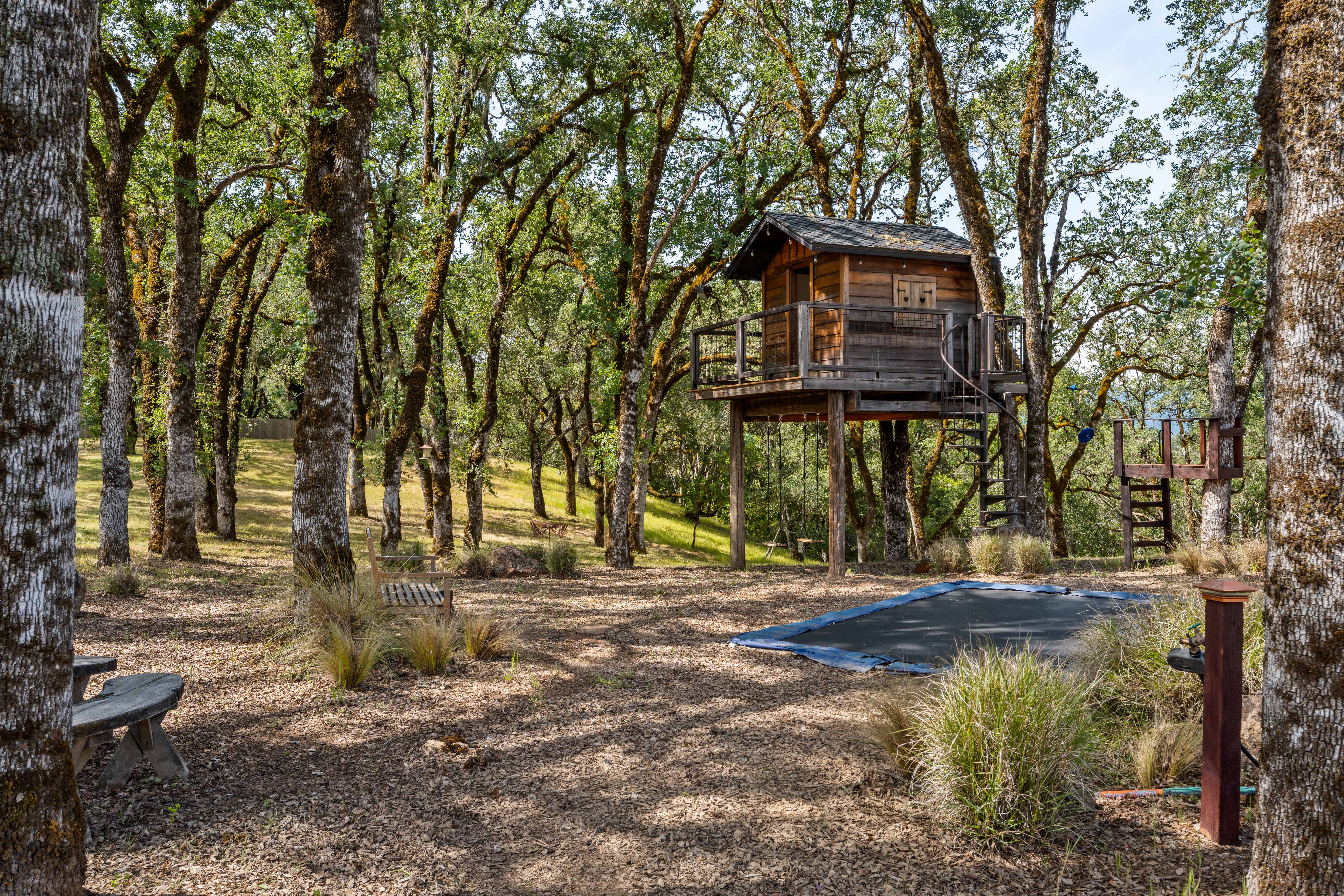 This serene backyard setting features a charming wooden treehouse elevated on stilts, complete with a deck and spiral staircase. The surrounding landscape is filled with mature oak trees, a sunken trampoline, and rustic wooden benches, creating a playful and tranquil outdoor retreat. The perspective is a wide-angle ground-level shot that captures the natural, wooded atmosphere of the property.