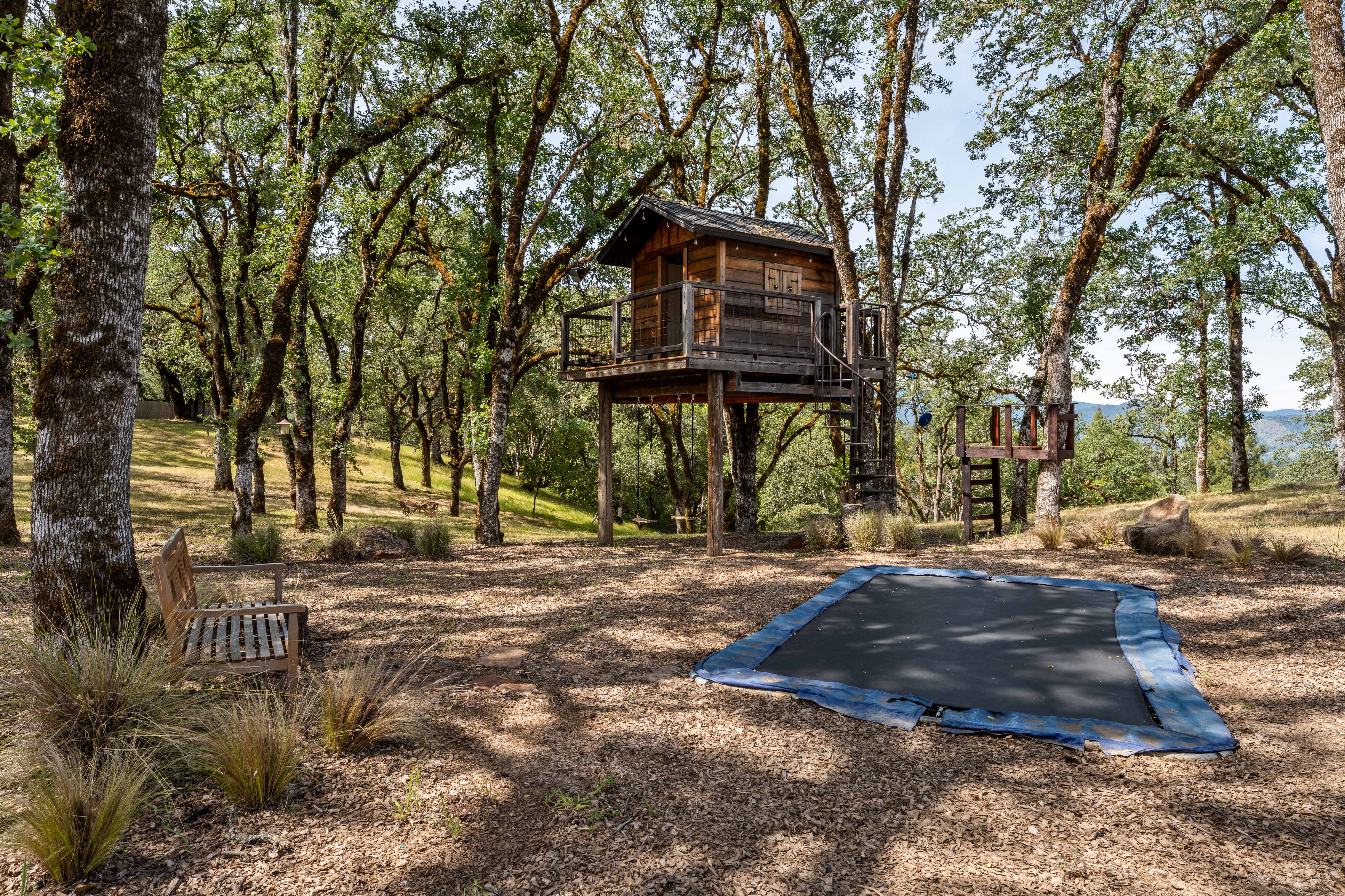 This serene outdoor space features a rustic wooden treehouse elevated on stilts, complete with a deck and spiral staircase, nestled among mature oak trees. In the foreground, a sunken in-ground trampoline sits on a wood-chipped ground, accompanied by a wooden bench, creating a playful and inviting backyard atmosphere. The scene is captured from a ground-level perspective, emphasizing the natural, wooded setting and the recreational amenities of the property.
