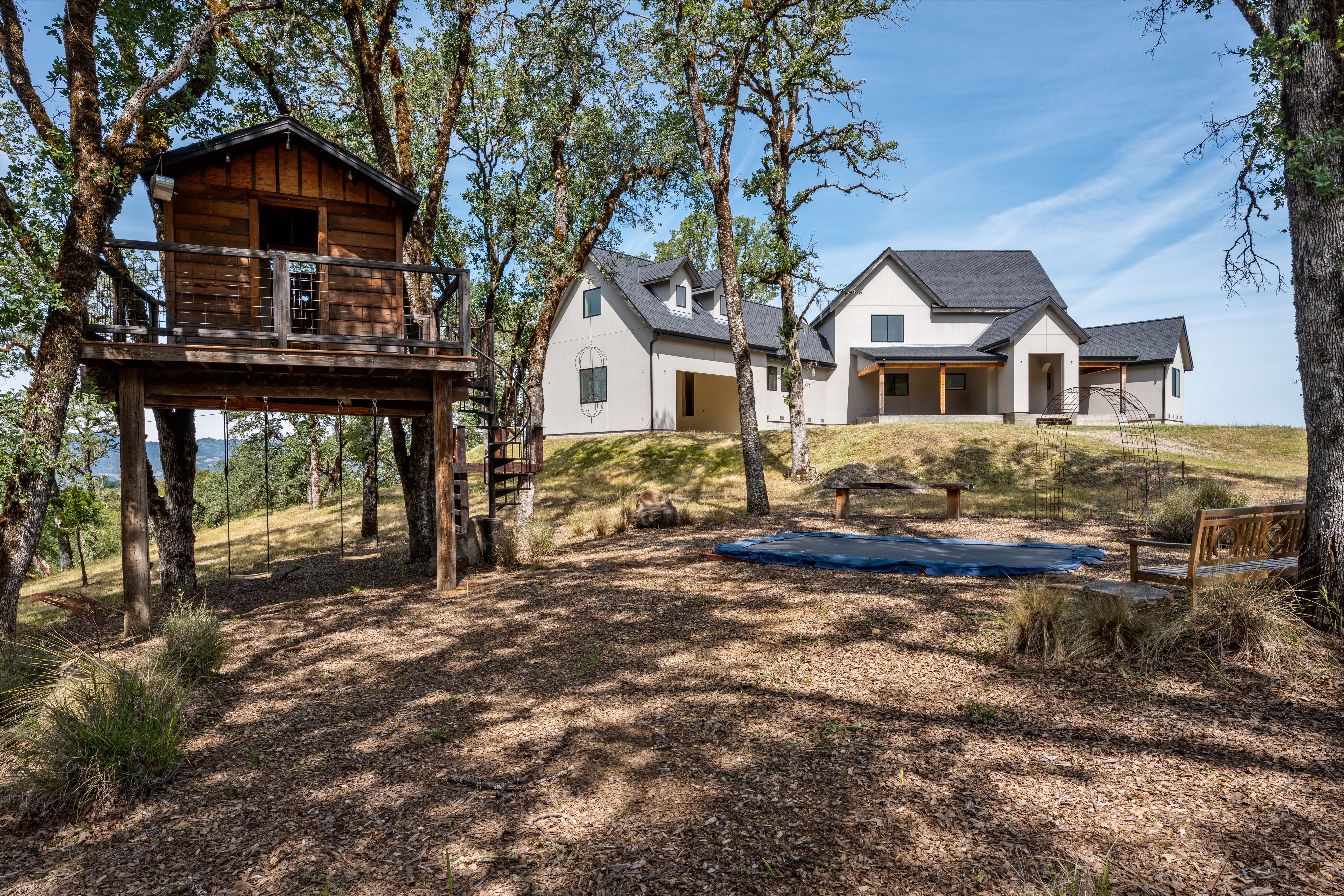 This scenic exterior view showcases a modern farmhouse-style home nestled on a grassy hill, surrounded by mature oak trees. In the foreground, a rustic wooden treehouse with a spiral staircase and attached swings provides a charming play area, while a sunken trampoline and a wooden bench offer additional outdoor recreation space. The composition captures a serene, rural lifestyle with a blend of architectural elegance and natural, wooded surroundings.